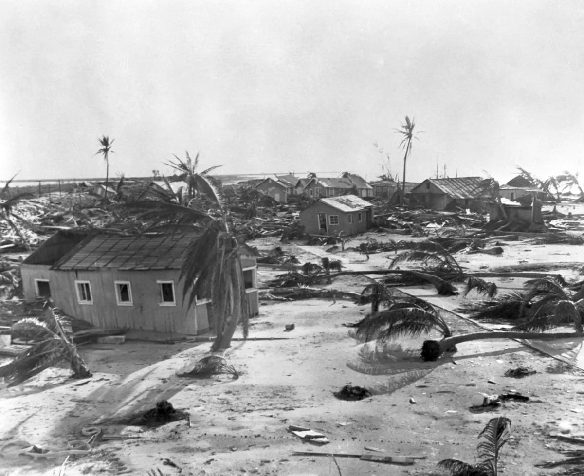 A black-and-white photograph of wrecked wooden houses, fallen palm trees and scattered debris on flat, sandy ground