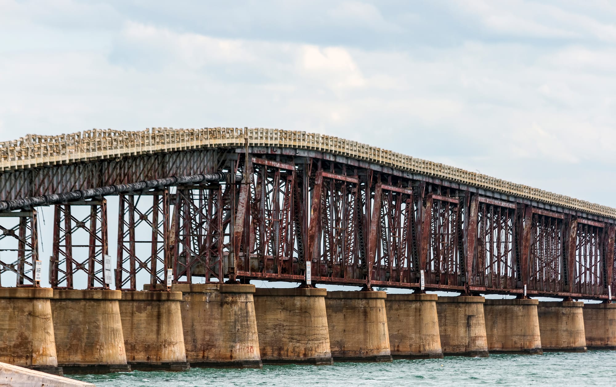 A long rusting steel truss bridge on concrete piers over the sea, with a concrete roadway and railings running along its top
