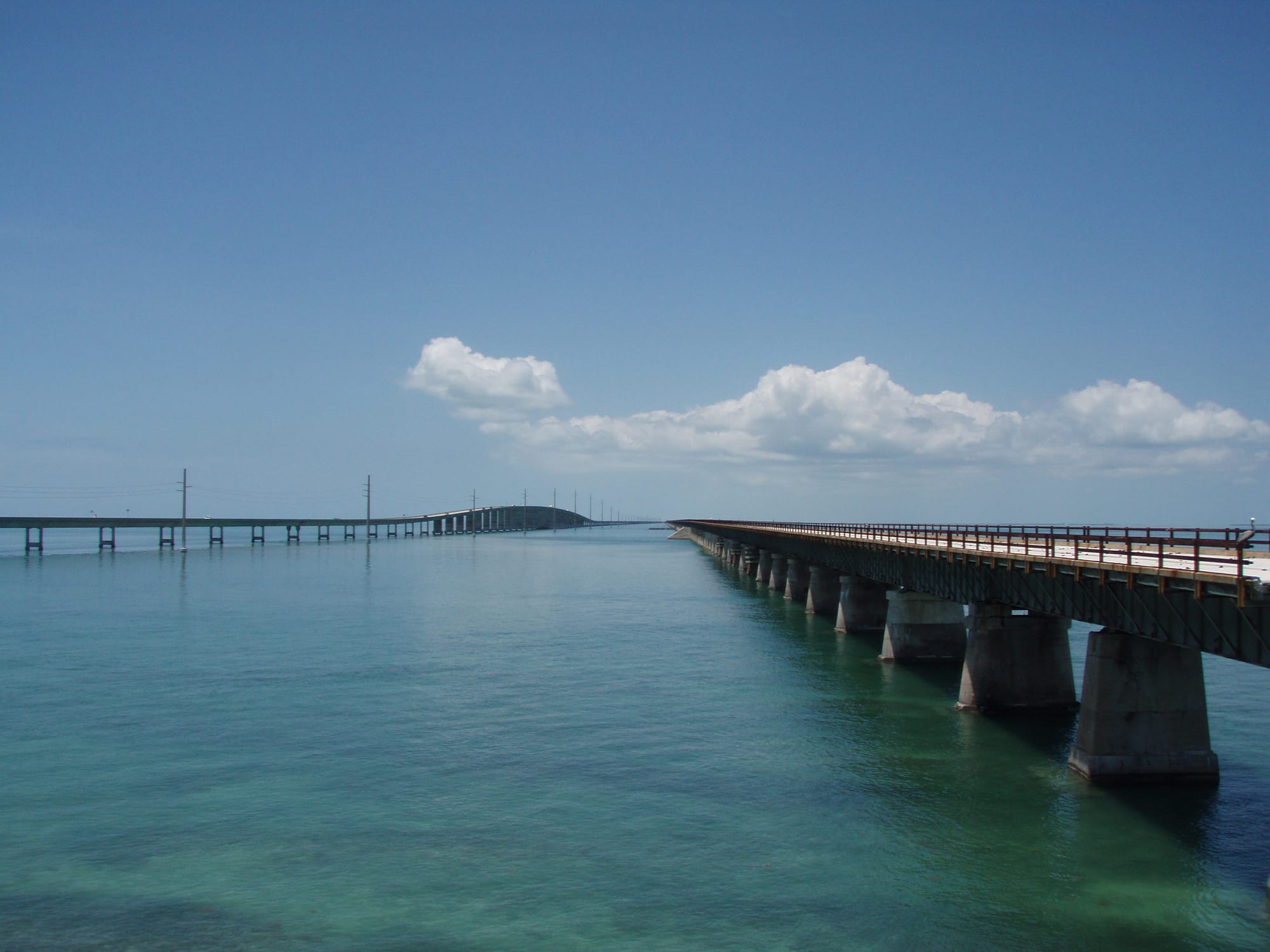 Two long, low bridges running side by side across calm turquoise water towards the horizon, the nearer one an older girder bridge on concrete piers
