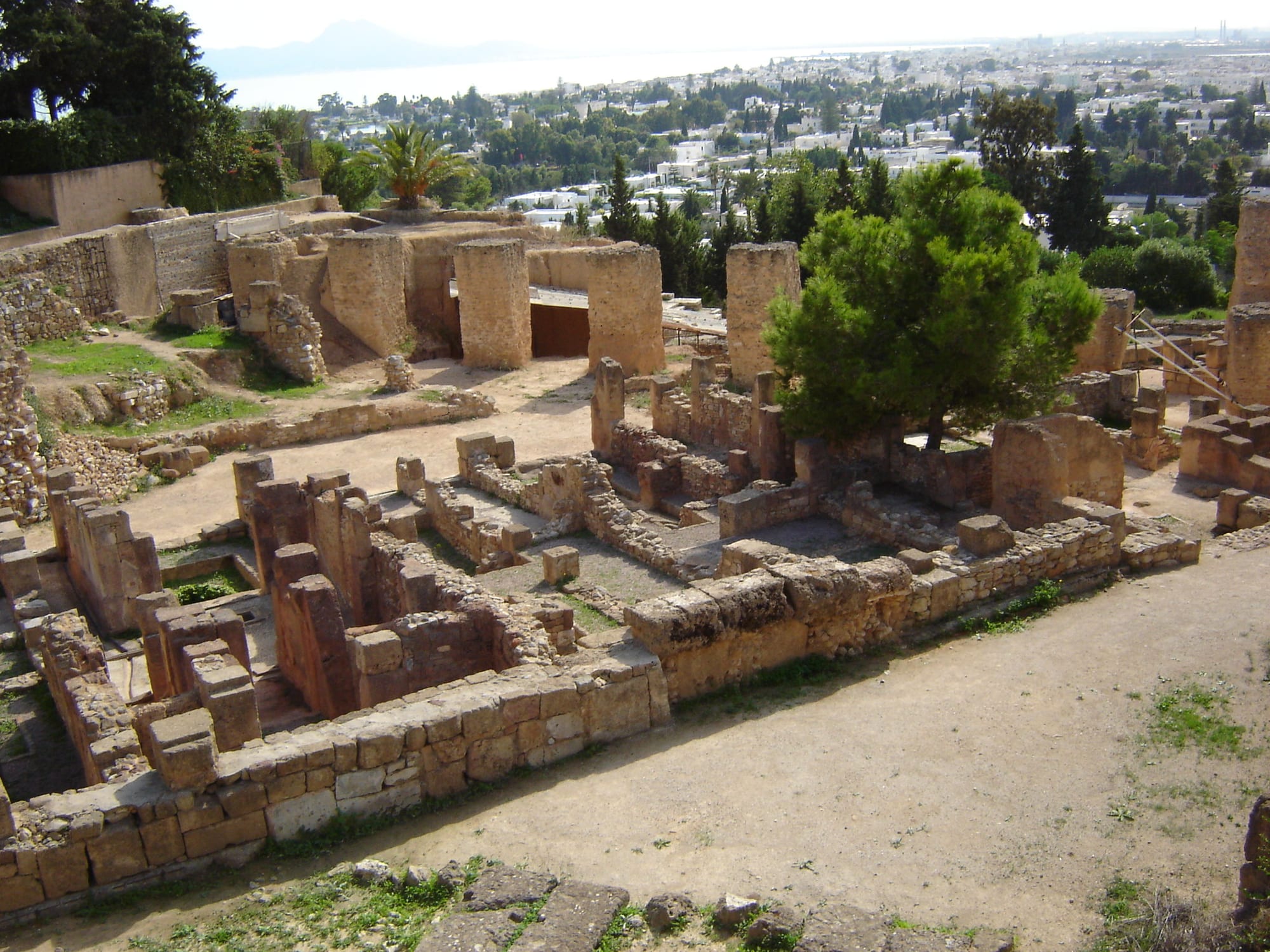 Excavated stone walls of ancient houses and streets on a hillside, with a modern town and the sea behind