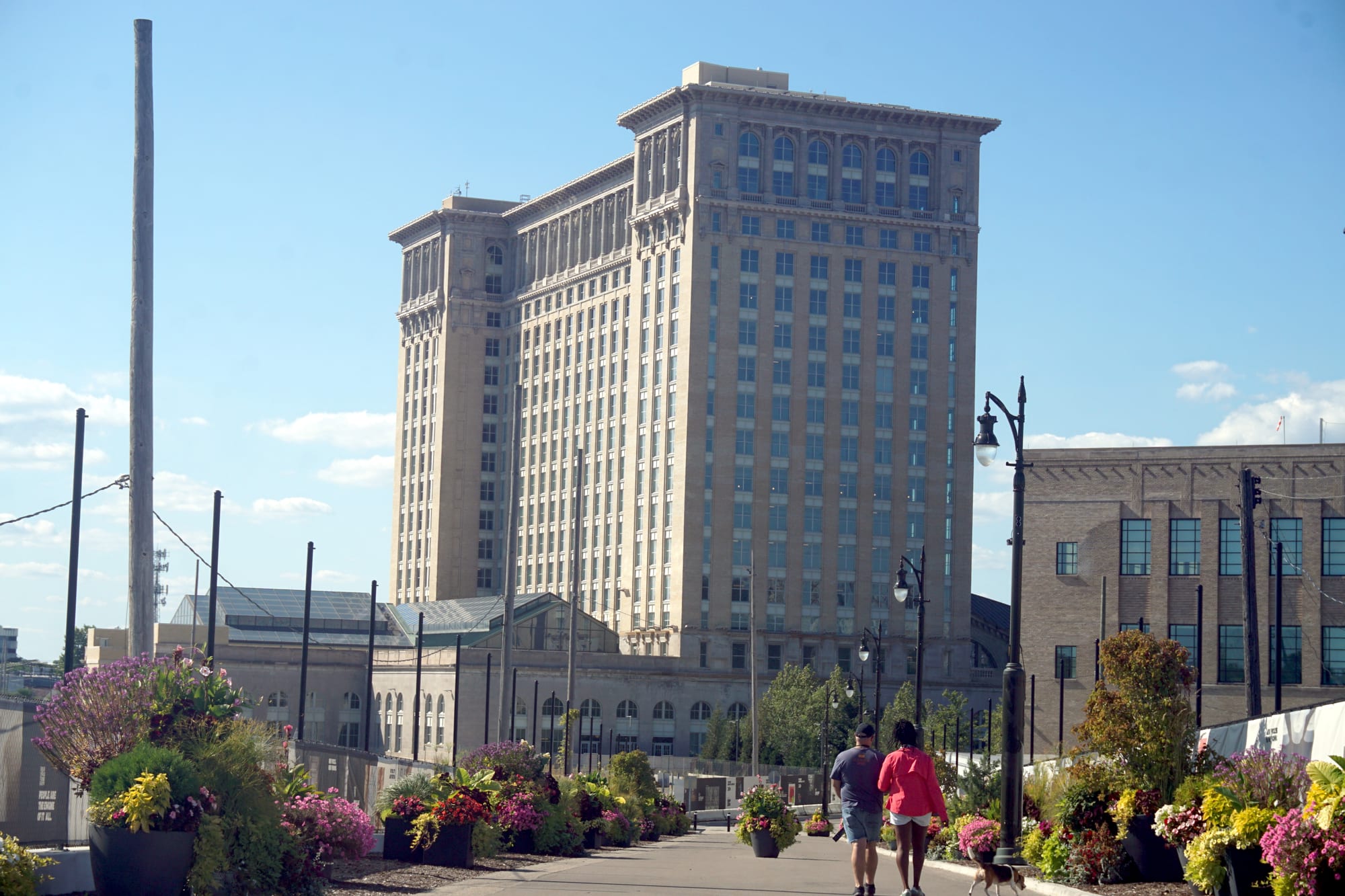 A restored stone railway station tower with clean windows behind a landscaped walkway lined with flower planters and people walking a dog