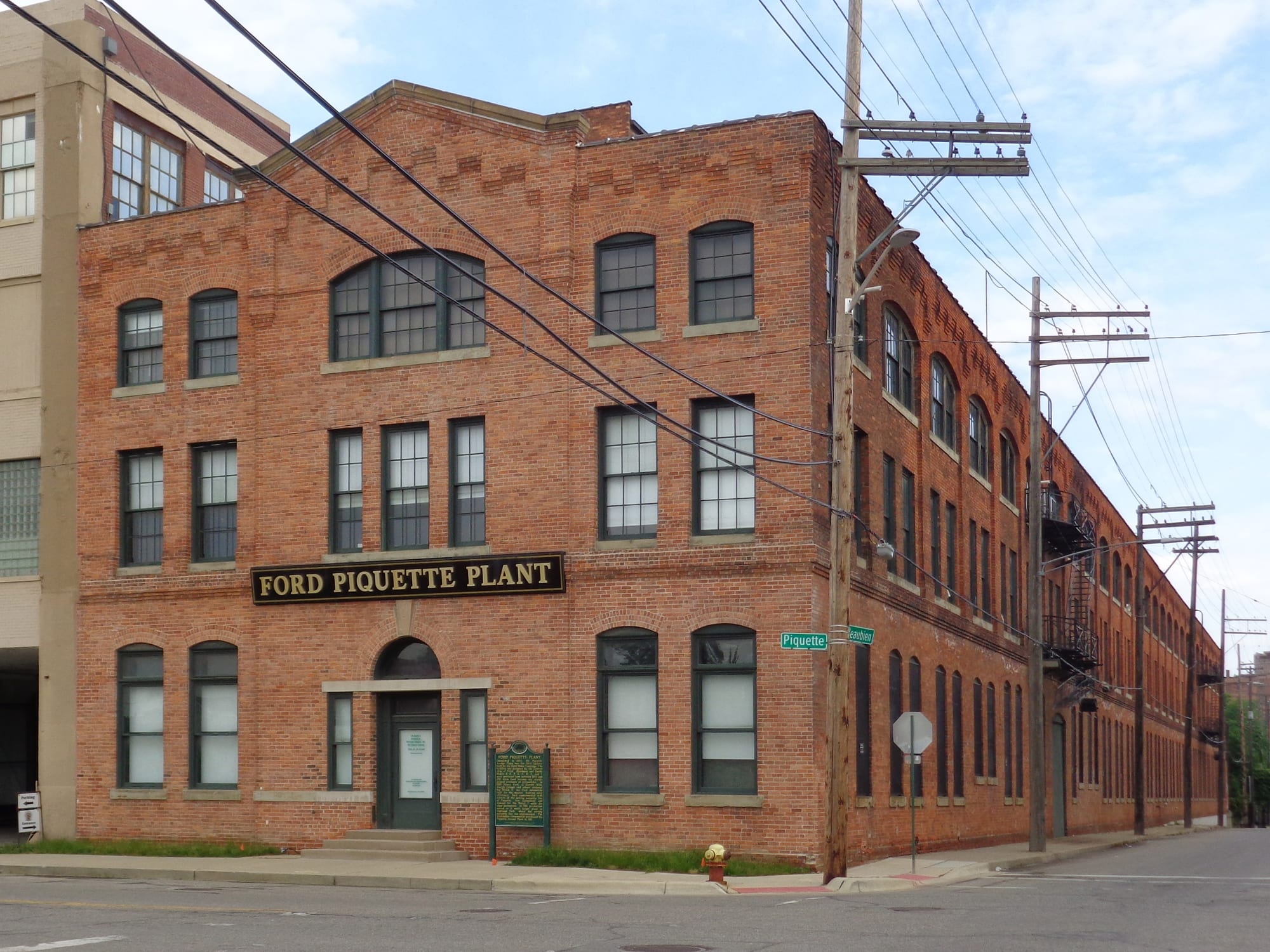 A three-storey red-brick factory with tall arched windows and a sign reading Ford Piquette Plant, on a street corner