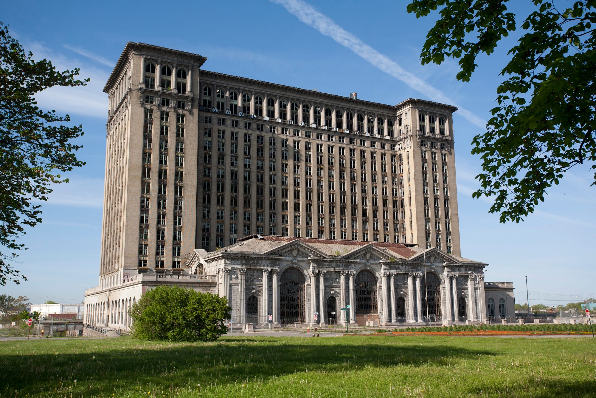 A huge abandoned railway station with a tall office tower, rows of empty broken windows and a columned entrance hall, seen across a lawn