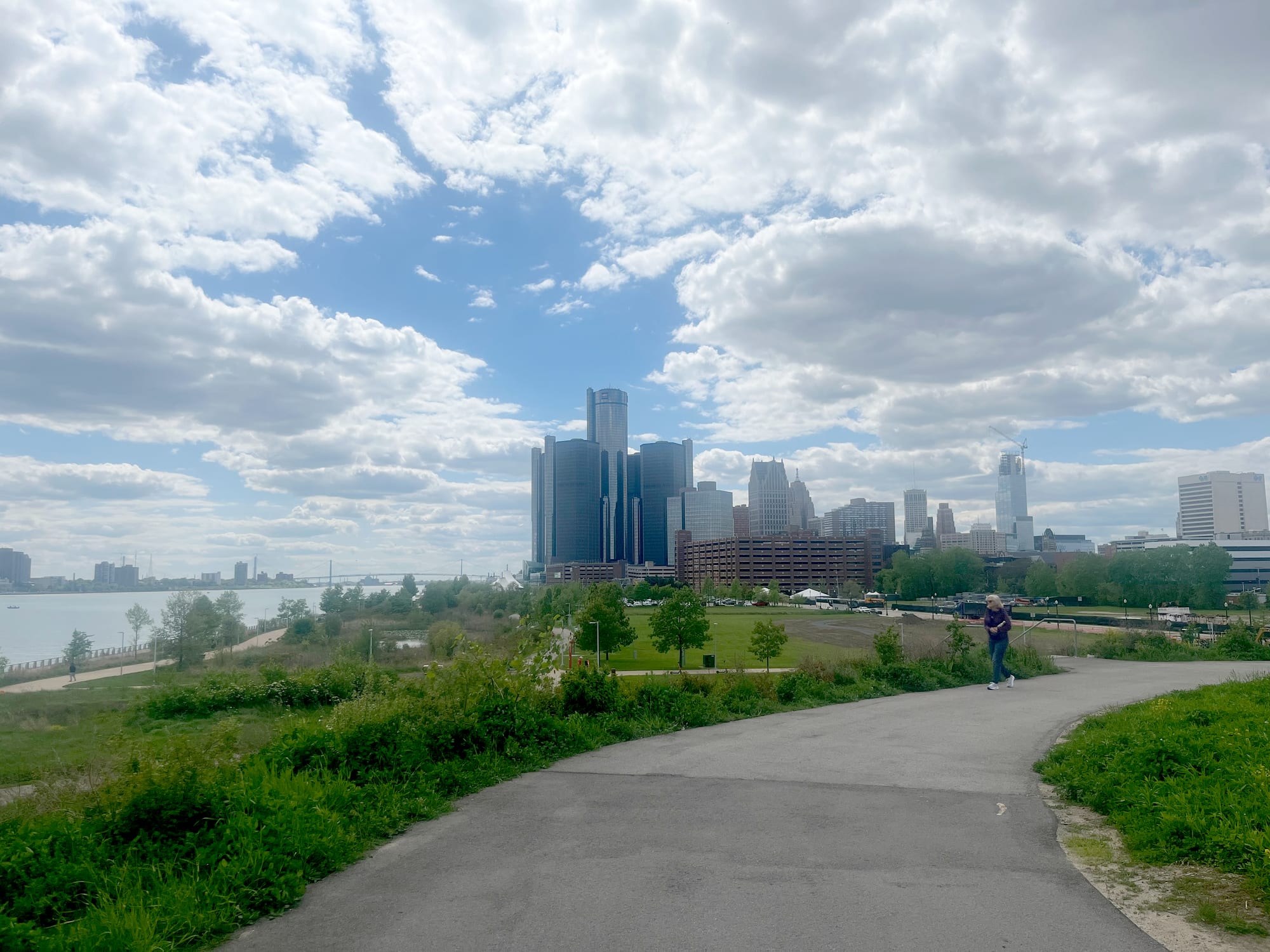 A riverside footpath and green park leading towards a cluster of dark glass towers and older skyscrapers under a cloudy sky