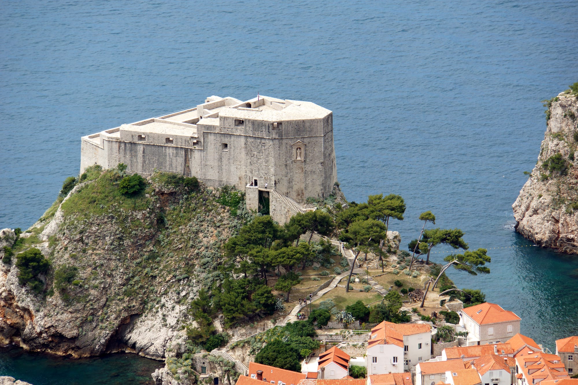 A pale stone fortress on top of a rocky, tree-covered headland above the deep blue sea, with orange roofs below