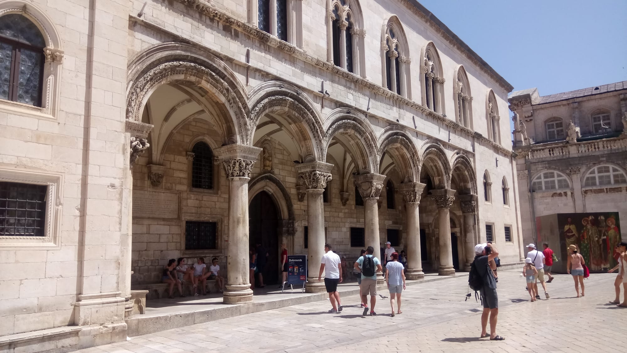 A pale stone palace with an arcade of carved columns and pointed upper windows, with visitors walking past on a sunny square