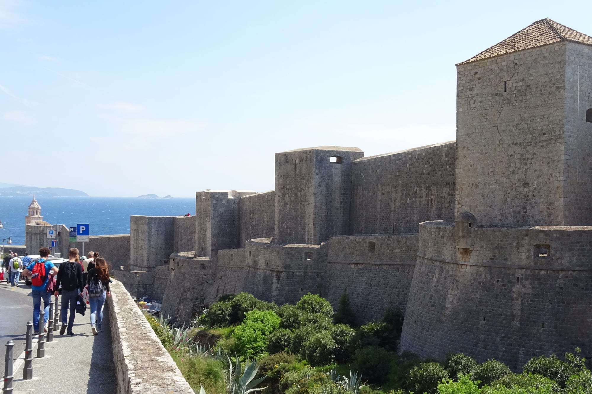 Massive grey stone walls and square towers rising above green shrubs, with walkers on a path beside them and the blue sea beyond