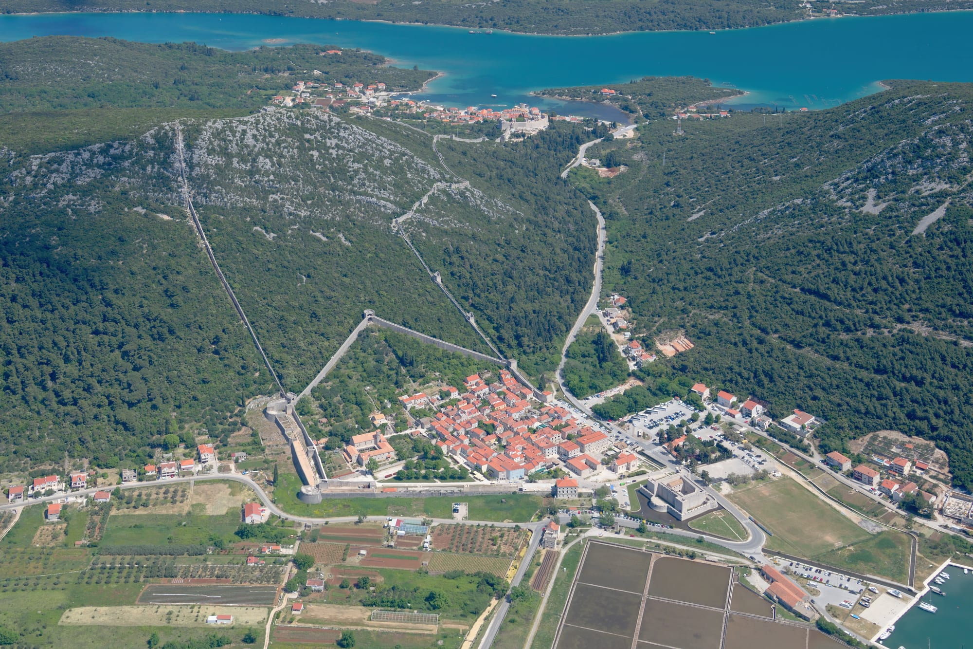 An aerial view of a small town of orange roofs with long stone walls climbing a forested hill to another village on a bay, and rectangular salt pans in the foreground