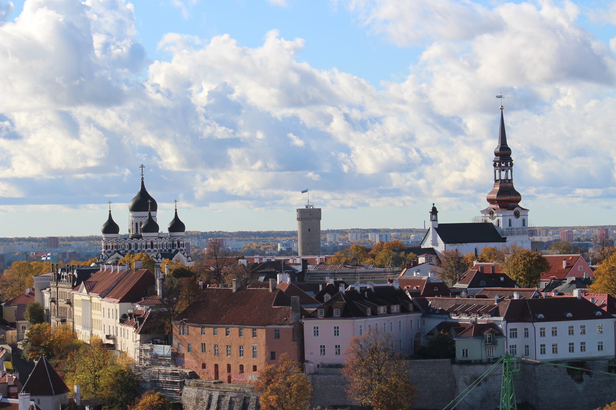 Red and orange roofs of an old town below a hill topped by an onion-domed cathedral, a round stone tower and a white church spire, under a cloudy blue sky
