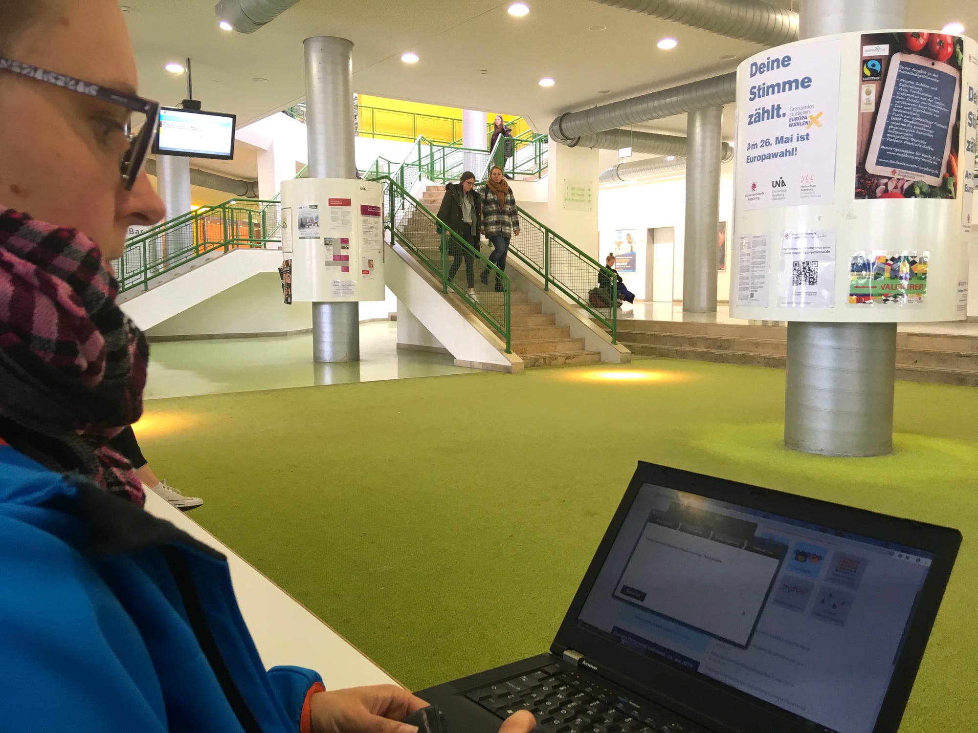 A woman in a scarf sits at a laptop in a university foyer with a green floor, stairs and posters about a European election