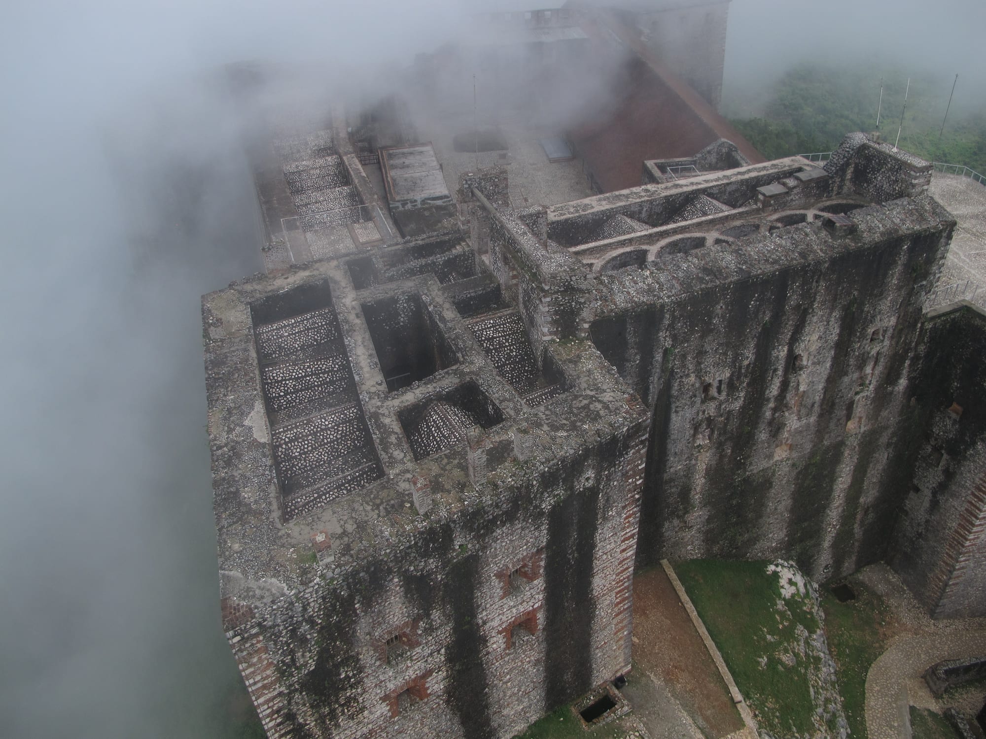 The massive grey stone walls and roof terraces of a mountaintop fortress seen from above, half hidden in cloud
