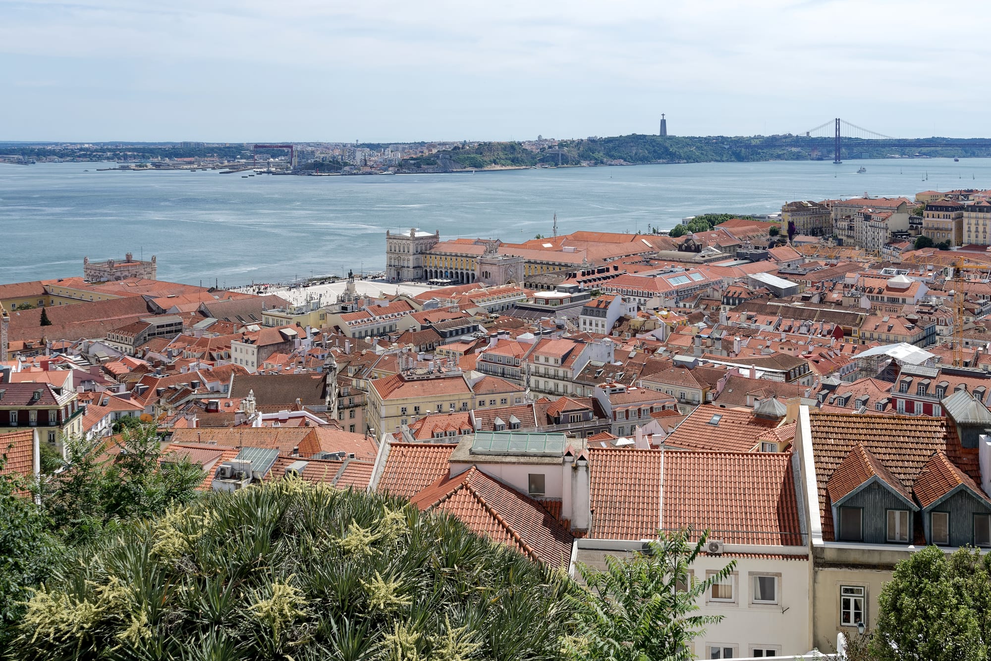 Red-tiled rooftops of a dense city district stretch down to a large arcaded square on the edge of a wide river, with a suspension bridge in the distance