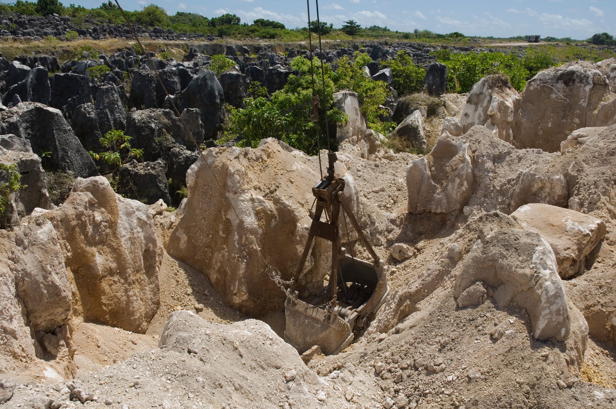 A rusty mechanical grab hanging from cables among pale limestone boulders and dark coral pinnacles