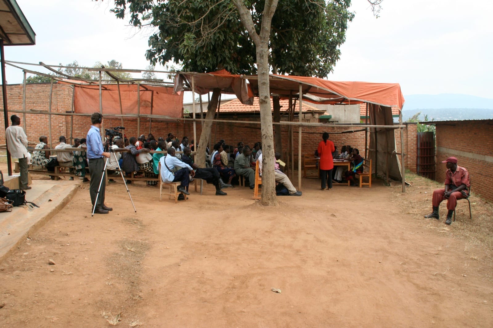 An open-air hearing under a tree and a makeshift shelter of poles and orange sheeting, with a seated crowd, a woman in red standing before a table of judges, and a man filming on a tripod