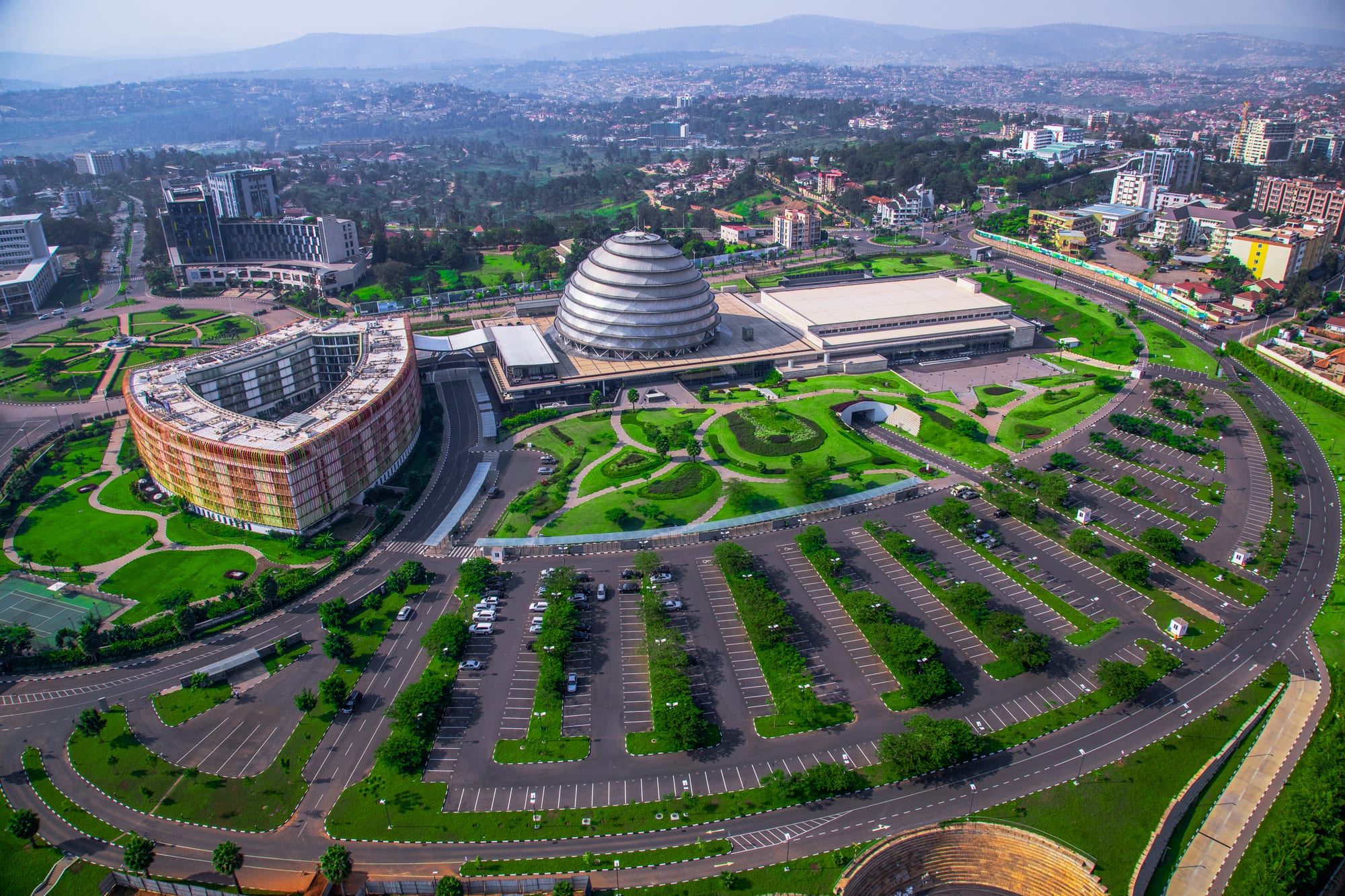 An aerial view of a modern city district with a large domed building, a curved multi-coloured hotel, landscaped gardens and car parks, with green hills behind