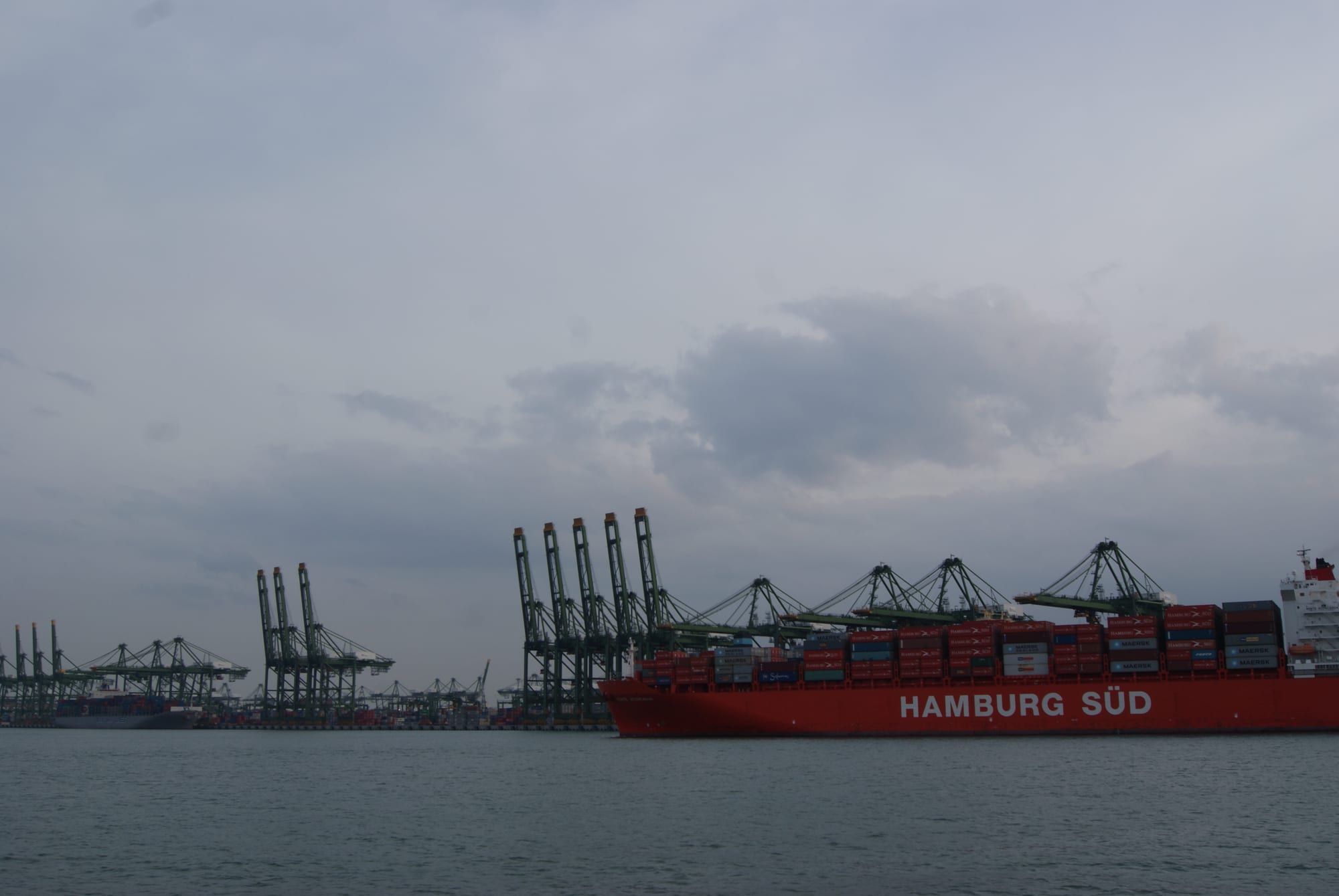 A red container ship moored beside a long row of green cranes at a port under a grey sky