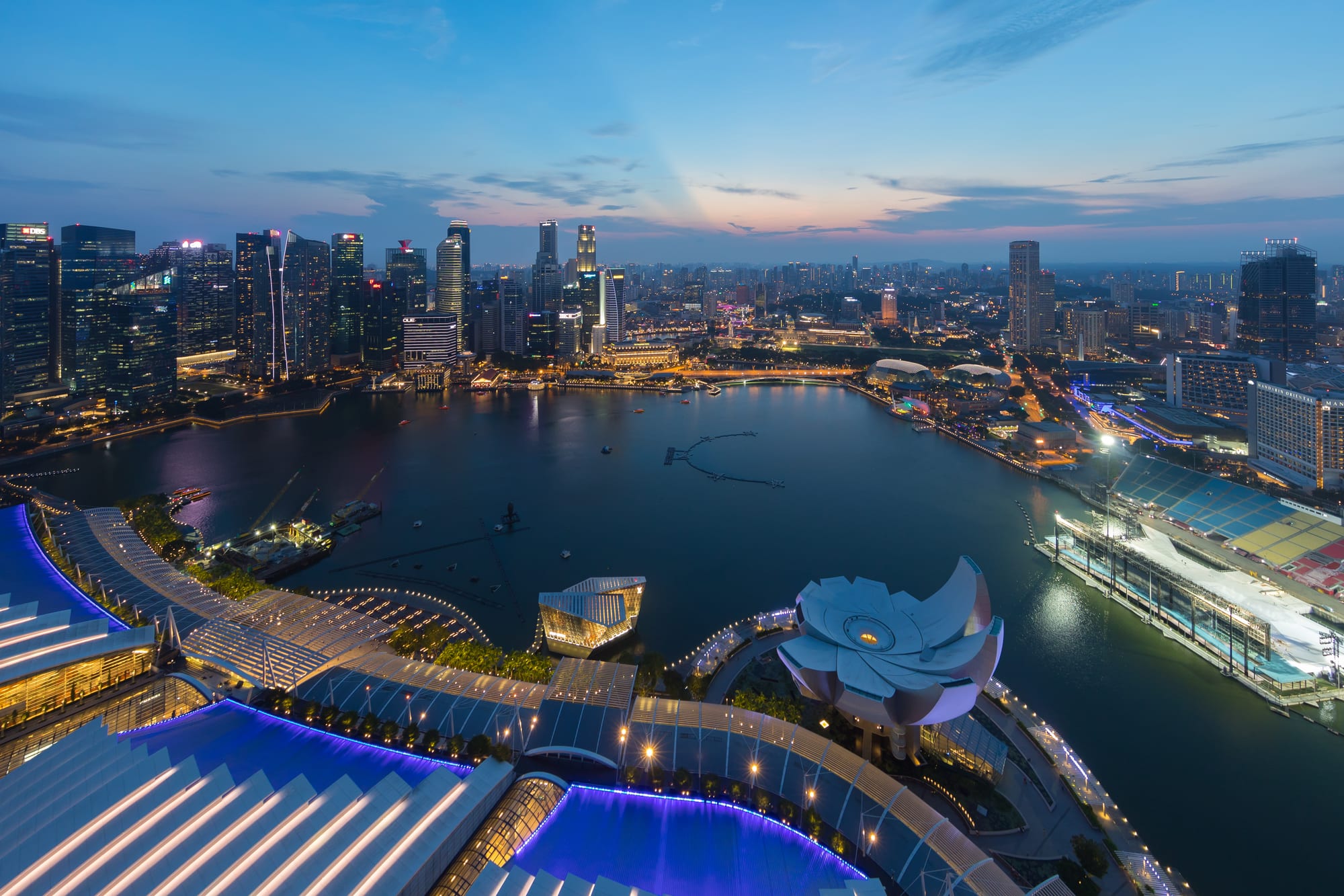 A curving bay ringed by lit skyscrapers at dusk, with a lotus-shaped museum and a floating platform in the foreground