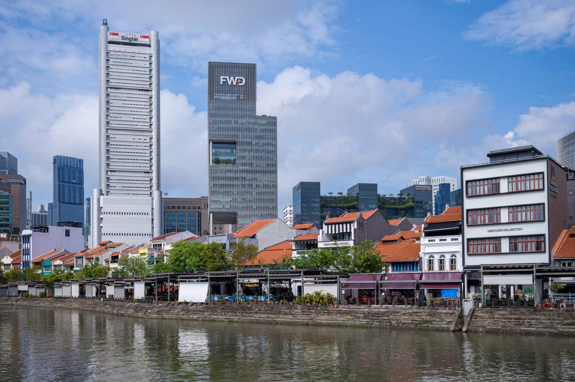A row of low old shophouses with red-tiled roofs along a river embankment, with tall office towers behind