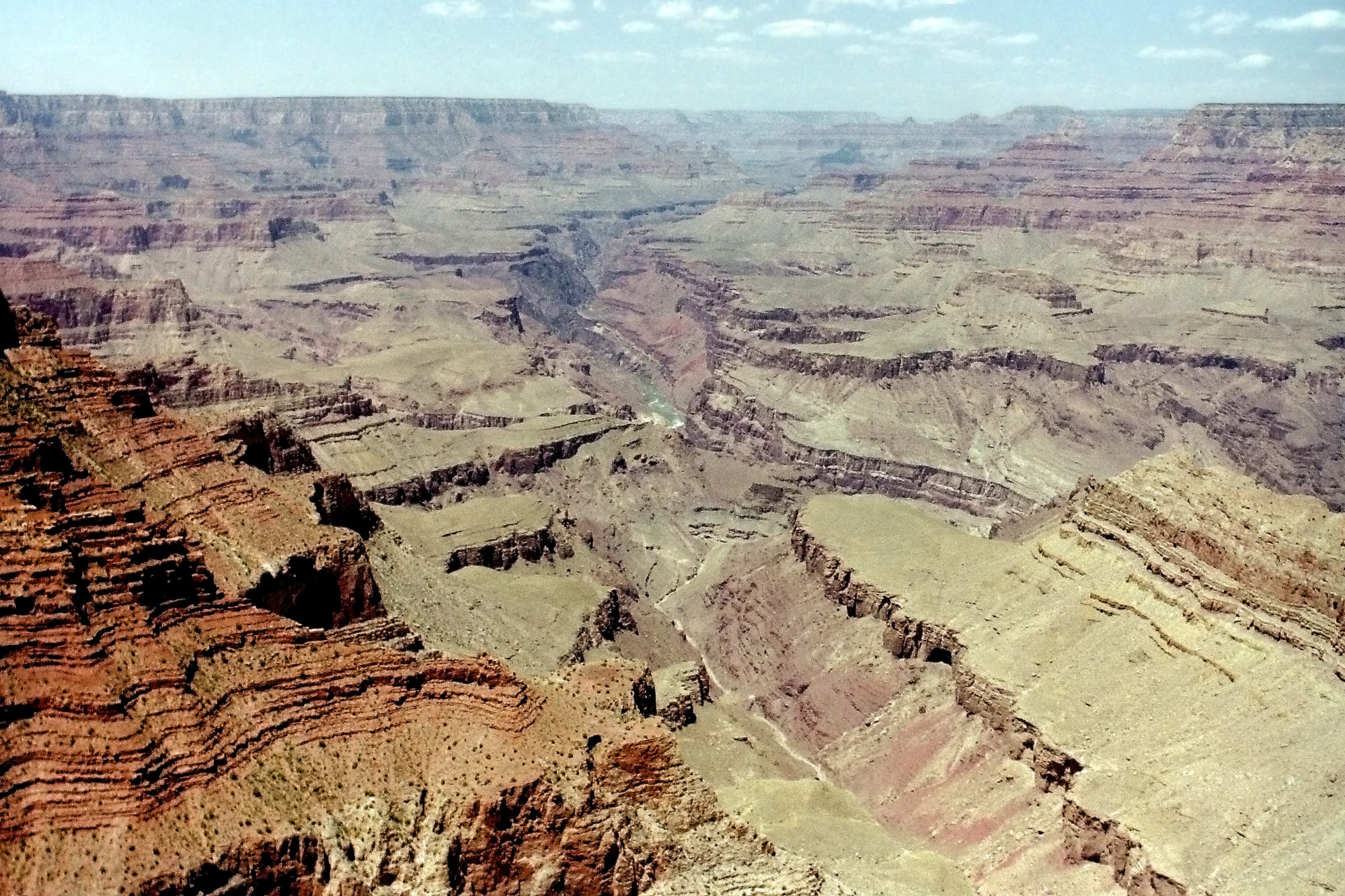 A vast canyon of red, tan and green rock terraces and cliffs stretching to a flat horizon, with a narrow green river at the bottom