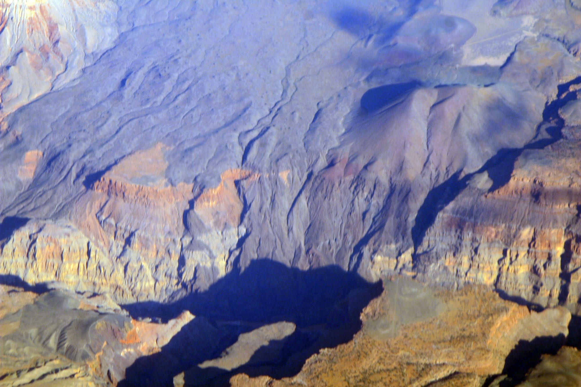 An aerial view of dark lava flows and a volcanic cone spilling over the layered rim of a deep canyon in low, bluish light