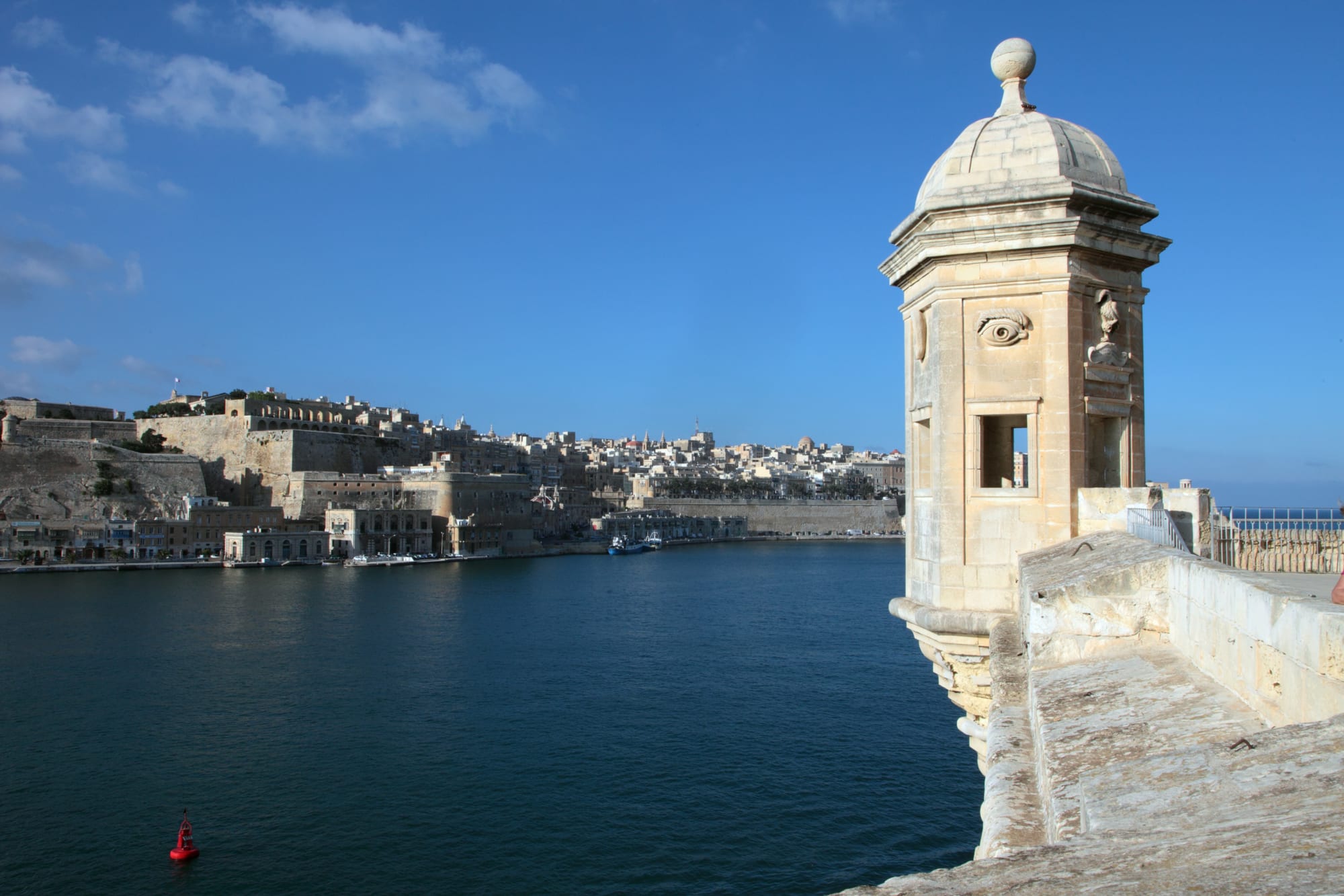 A carved stone sentry box on the corner of a bastion, looking across a deep blue harbour to a walled city of pale stone