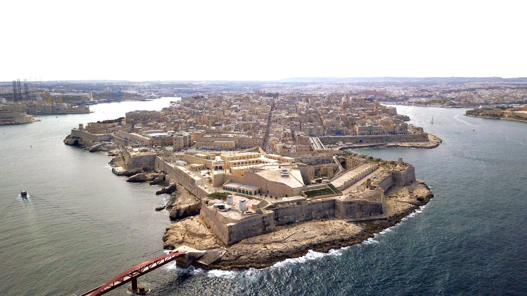 An aerial view of a walled city on a narrow peninsula between two harbours, with a pointed star-shaped fort at its tip and a red footbridge in the foreground