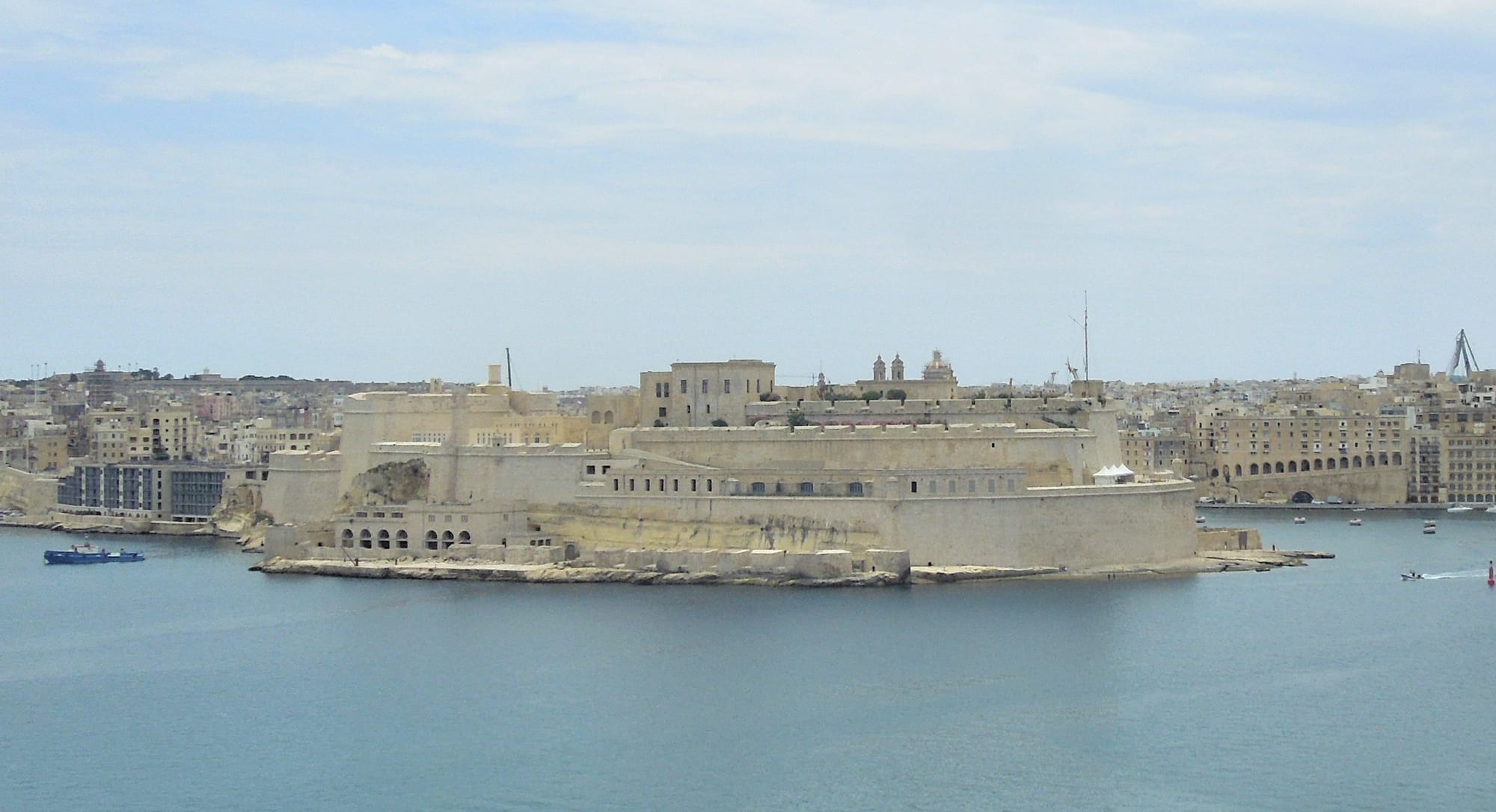 A large pale limestone fortress rising from the water at the tip of a town, seen across a calm blue harbour