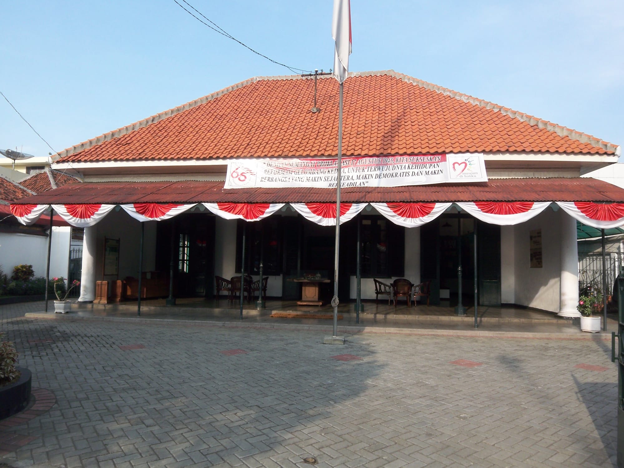 A low colonial-era house with a red tiled roof and a veranda hung with red and white bunting, behind a paved courtyard with a flagpole