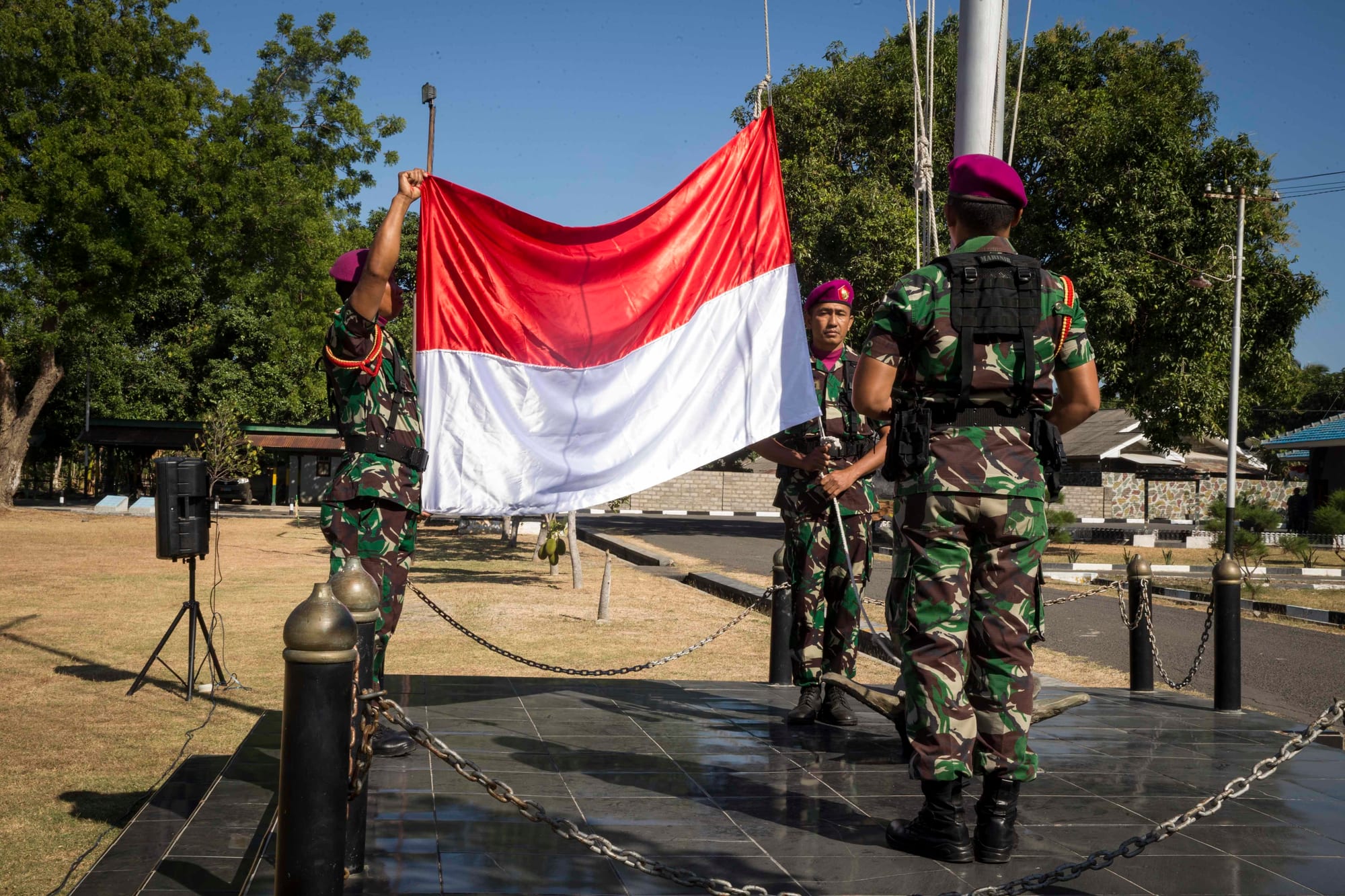 Soldiers in camouflage and maroon berets unfurling a large red and white flag beside a flagpole