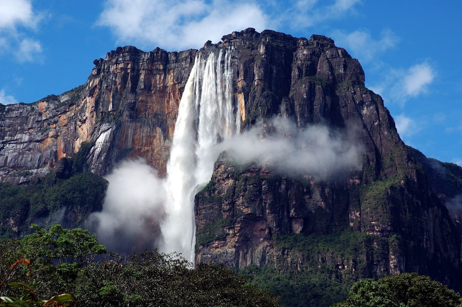 A thick white waterfall pouring from the top of a sheer, layered cliff of dark and orange rock, with spray drifting across the rock face and forest below