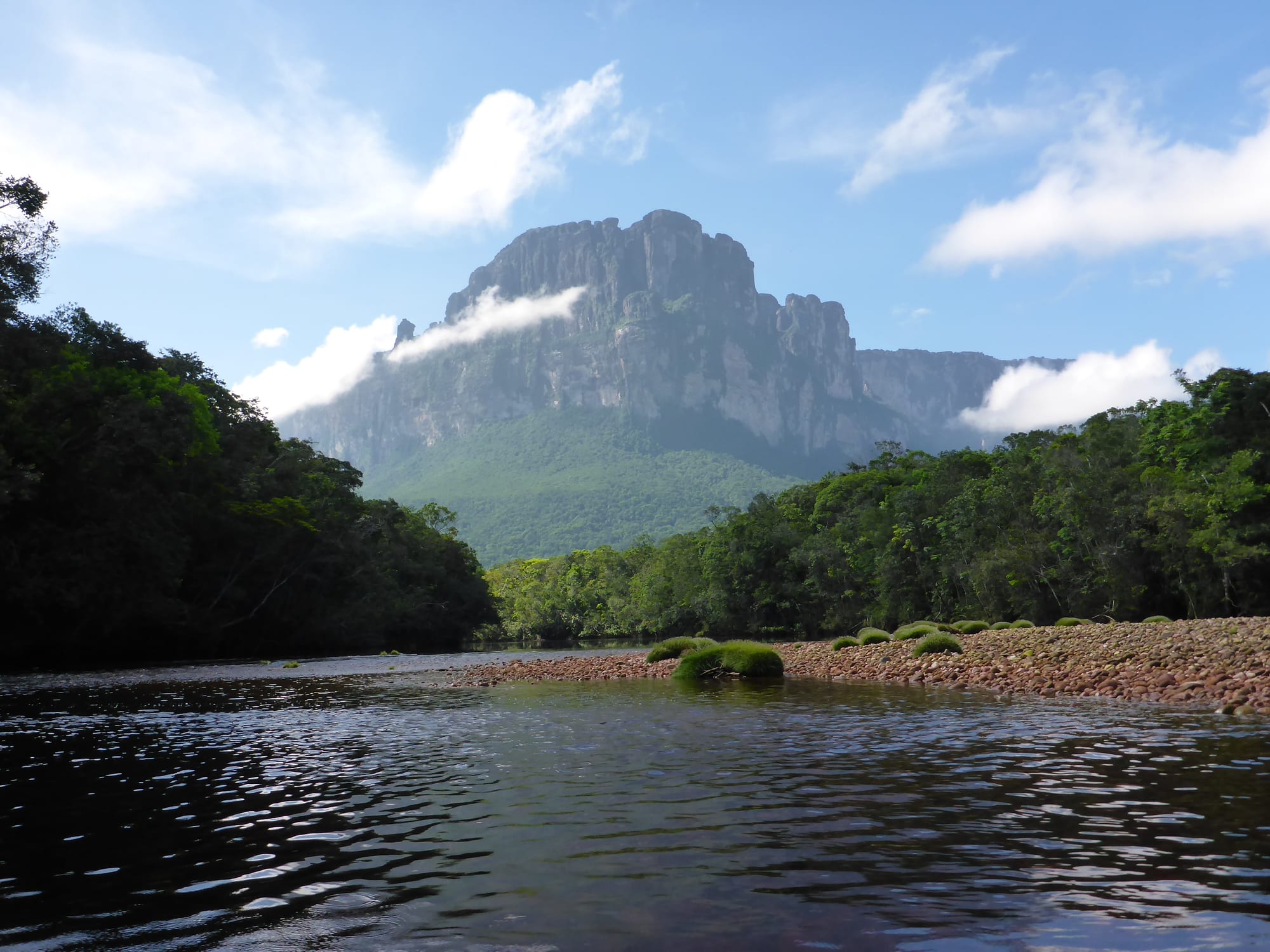 A dark river with a pebble bank in the foreground, forest on both sides and a towering flat-sided mountain under white cloud beyond