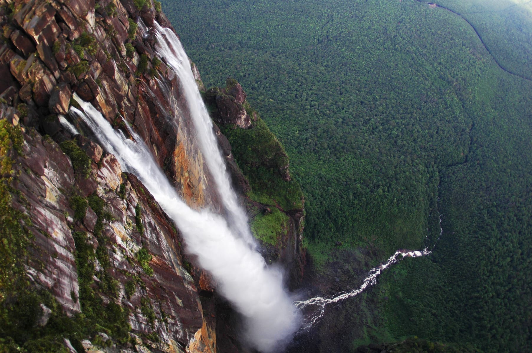 Looking down from the air along a waterfall falling down a red-brown cliff into a gorge, with a river winding through unbroken green forest below