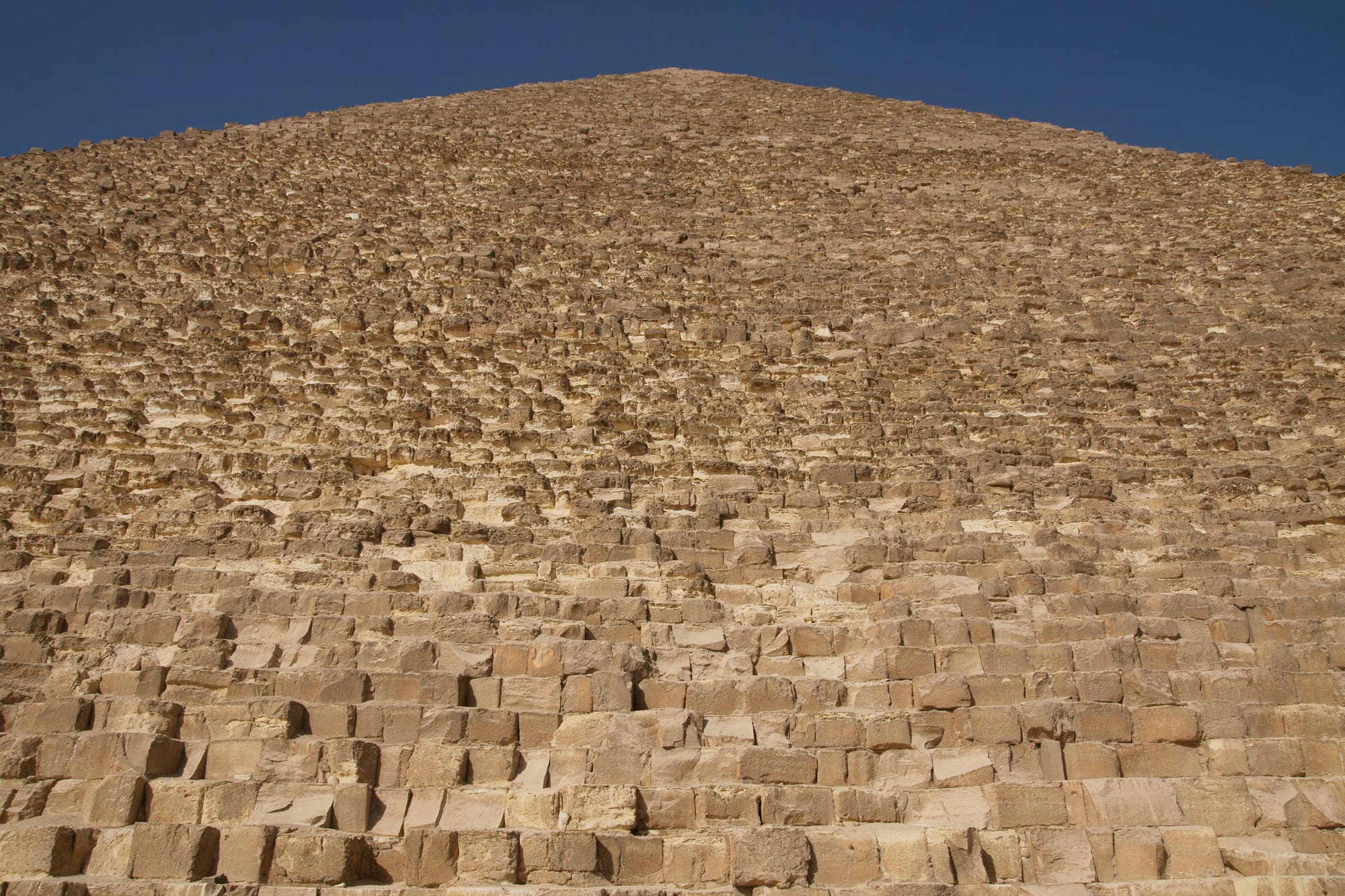 Rows of rough, weathered limestone blocks rising in steps towards the apex of a pyramid against a deep blue sky