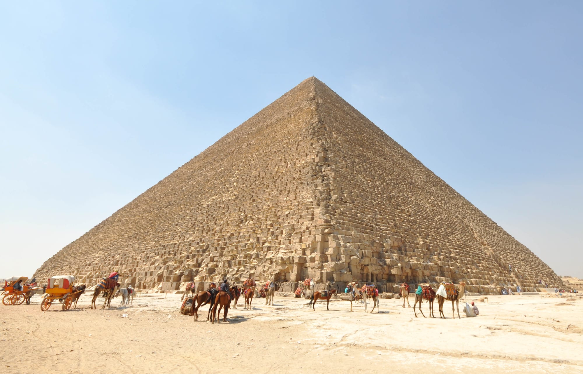 A huge stepped stone pyramid under a pale blue sky, with camels, horses and carriages waiting on the sand at its base