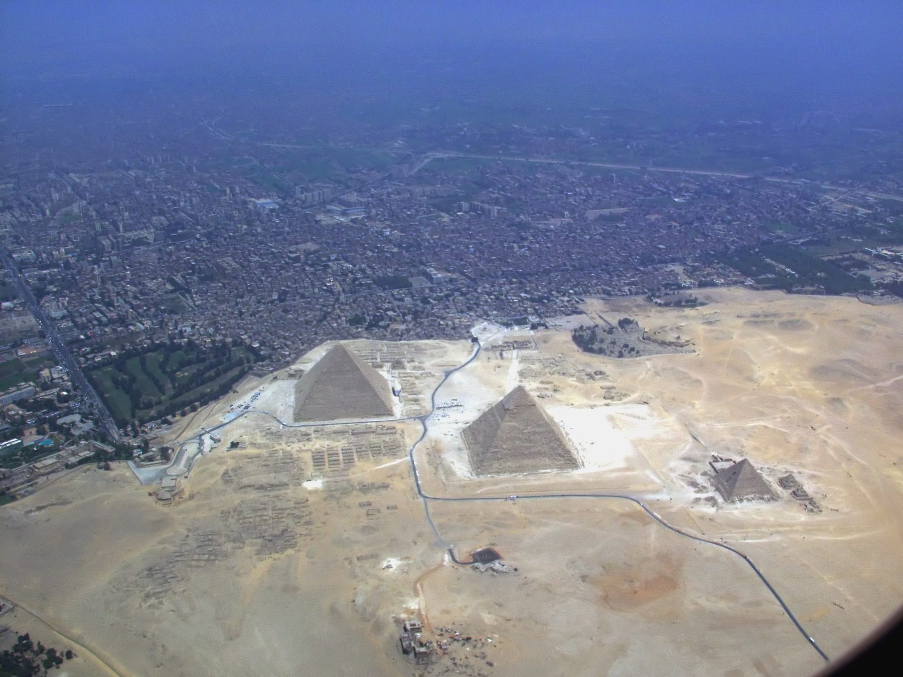 An aerial view of three pyramids on a sandy desert plateau, with rows of small tombs beside them and a dense city spreading right up to the desert edge