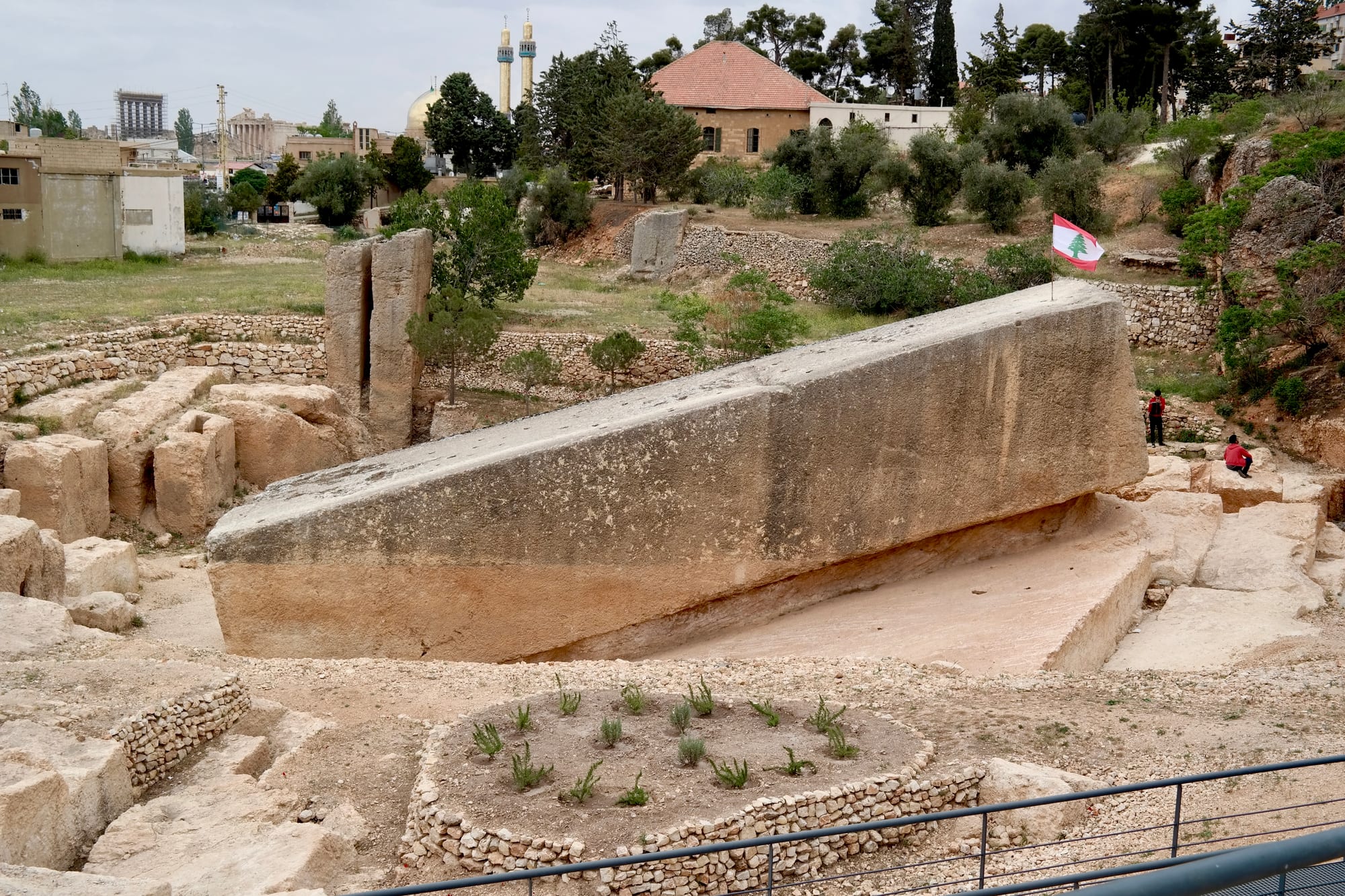 A gigantic rectangular block of pale limestone lying at a slant in an excavated quarry, with a Lebanese flag beside it and the town and a mosque's minarets behind