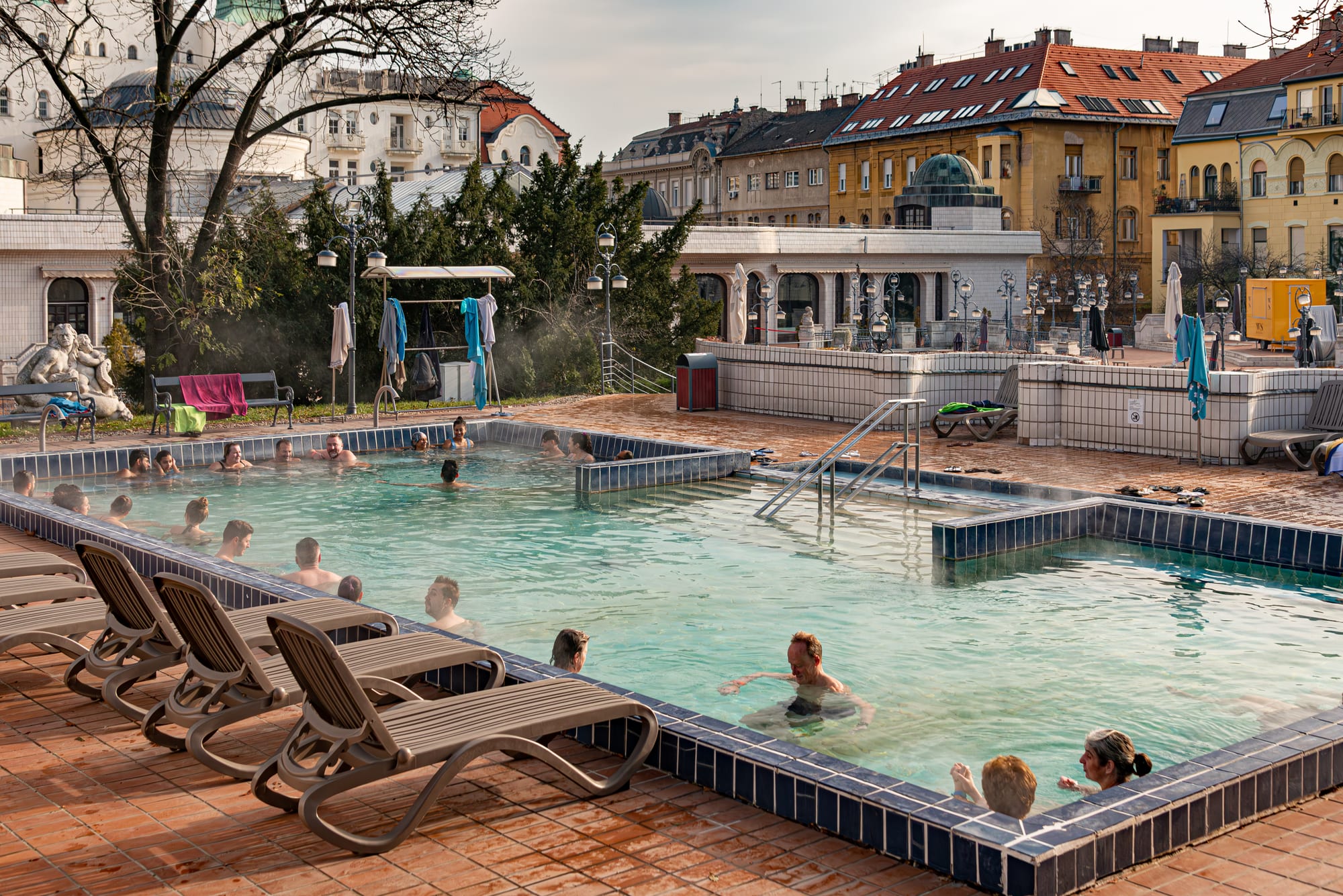 People bathing in a tiled outdoor thermal pool with sun loungers in the foreground and Budapest apartment buildings behind