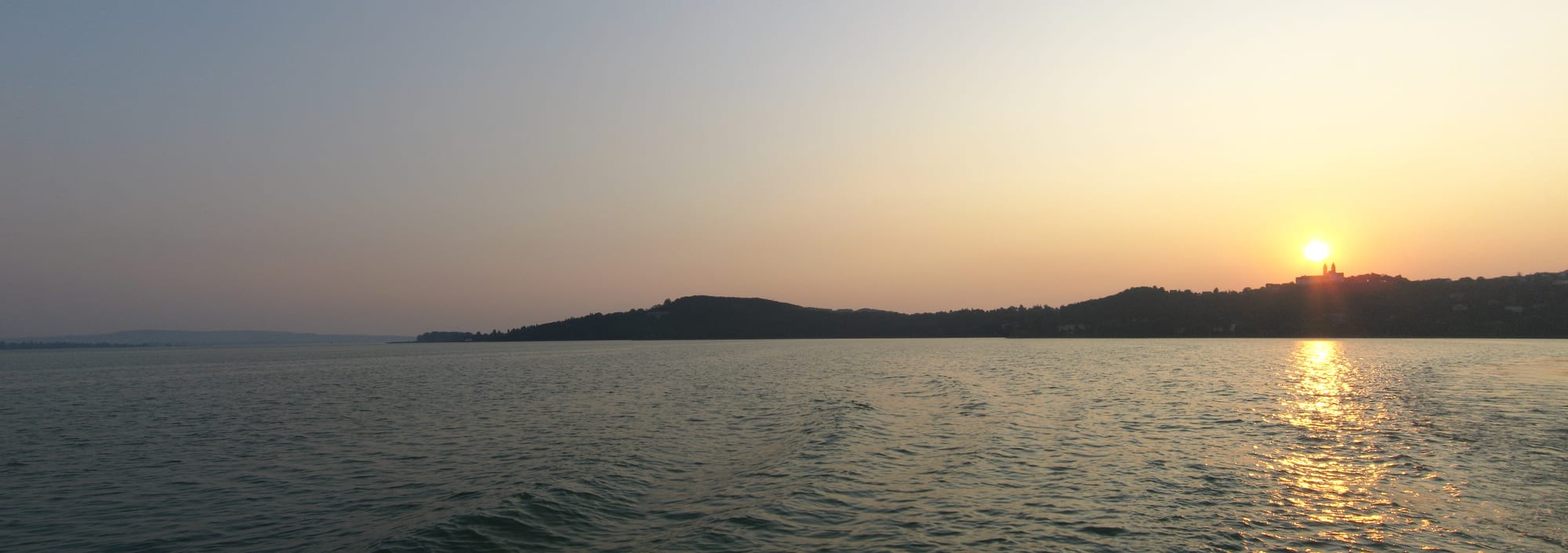 A twin-towered baroque abbey on a green peninsula above a wide calm lake at sunset