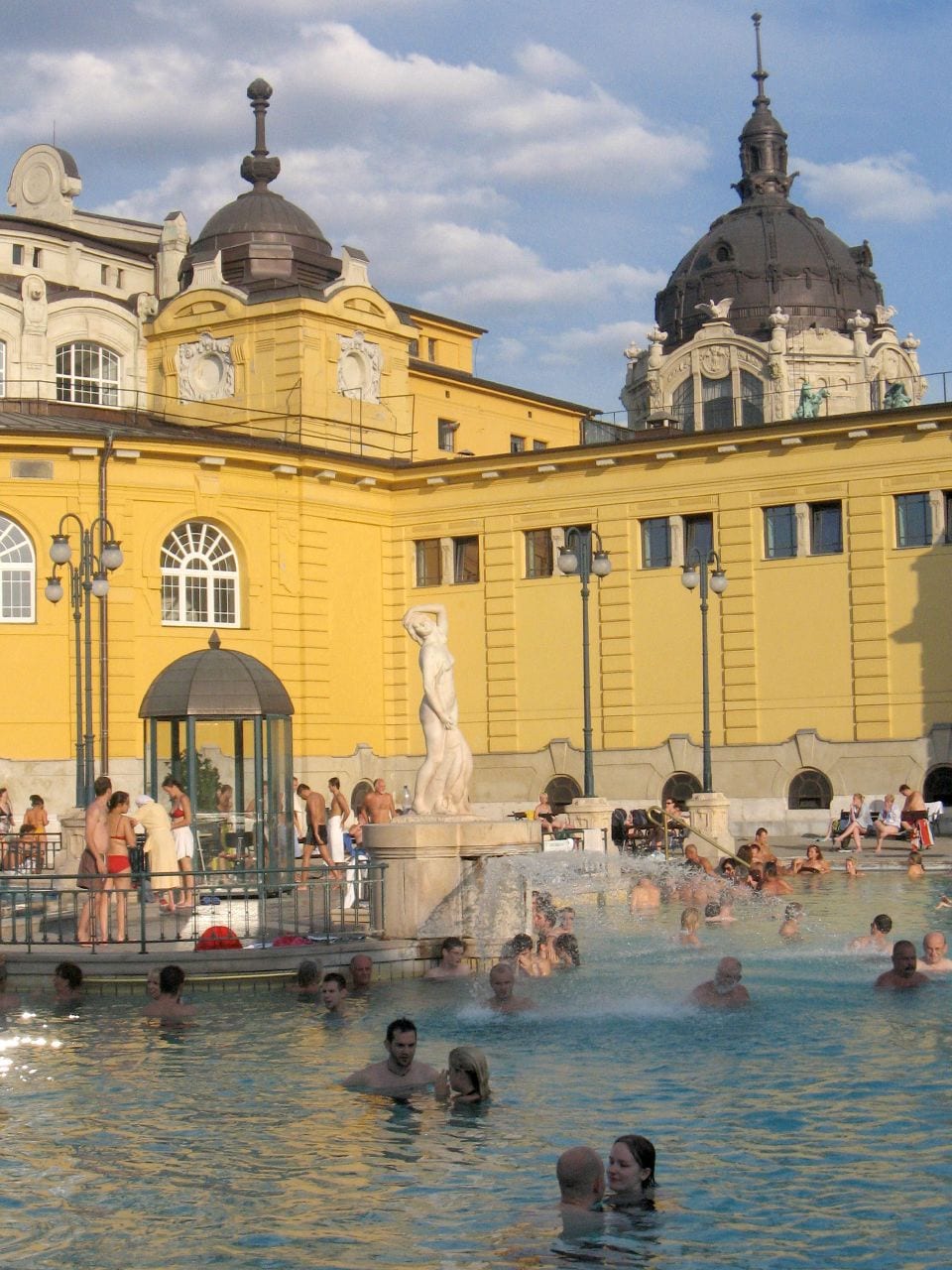 Bathers in a large outdoor thermal pool surrounded by a yellow neo-baroque palace