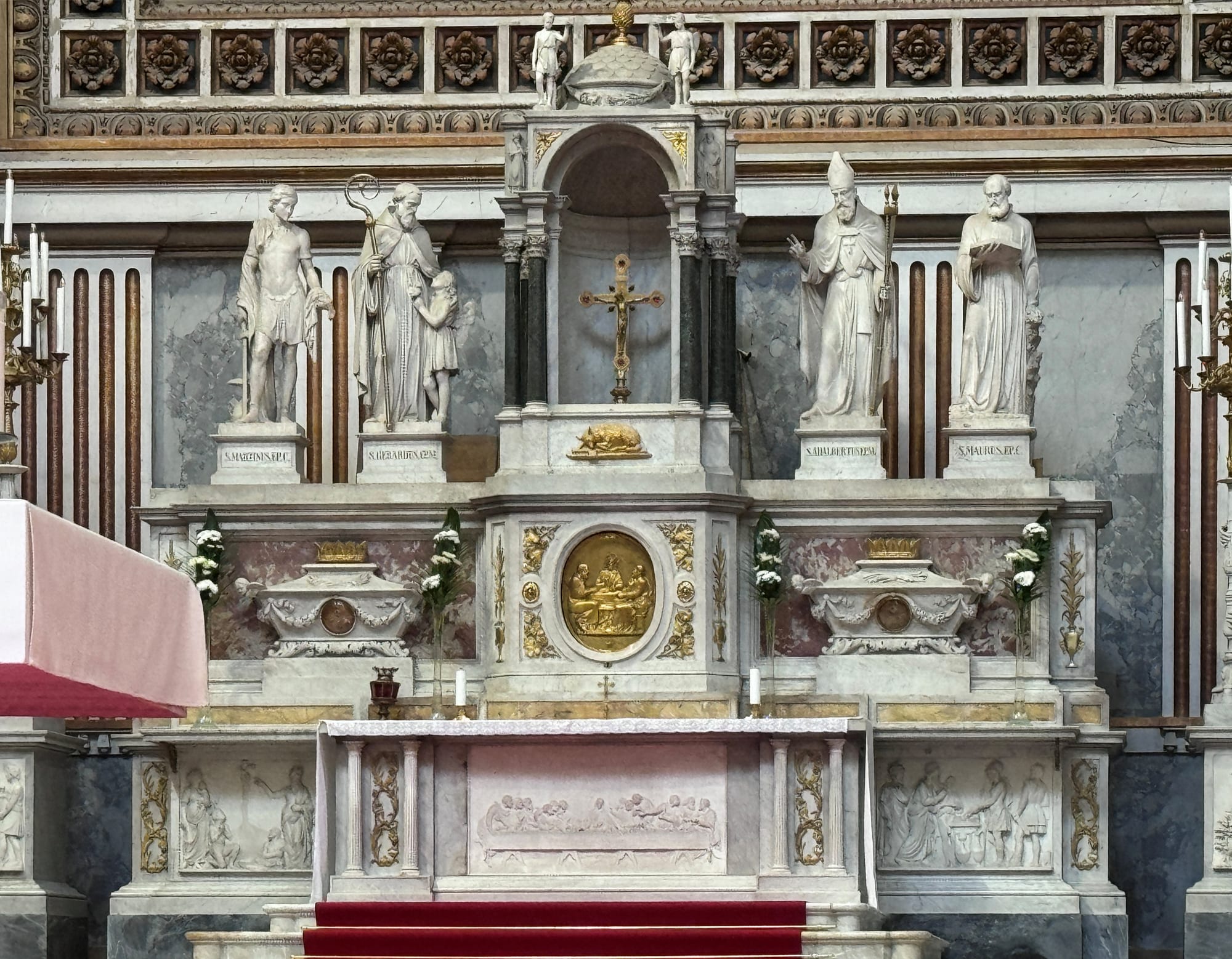 A white marble high altar with a gilded crucifix at its centre, flanked by four large statues of saints and bishops, with white flowers on the altar steps
