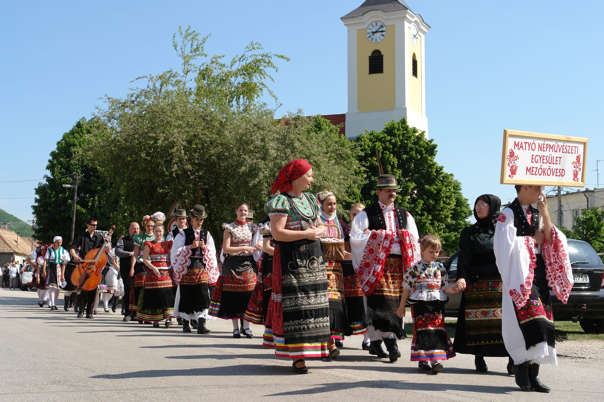 A procession of men and women in colourful folk costume with red floral embroidery walking along a village road behind a sign-bearer, with a musician carrying a double bass and a yellow church tower behind