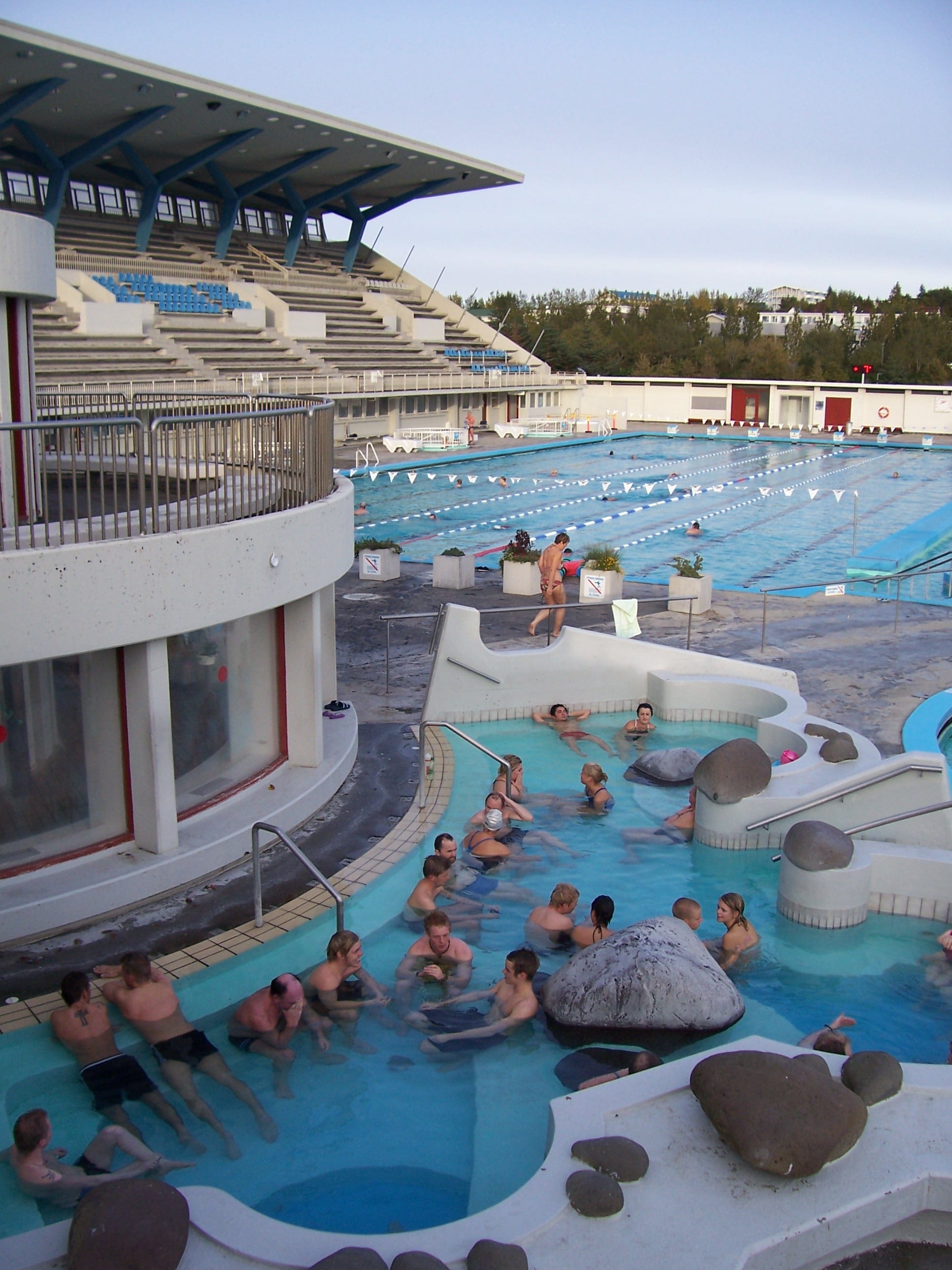 People soaking in outdoor hot pots at Laugardalslaug, a geothermal swimming pool complex in Reykjavík, with lap pool lanes behind