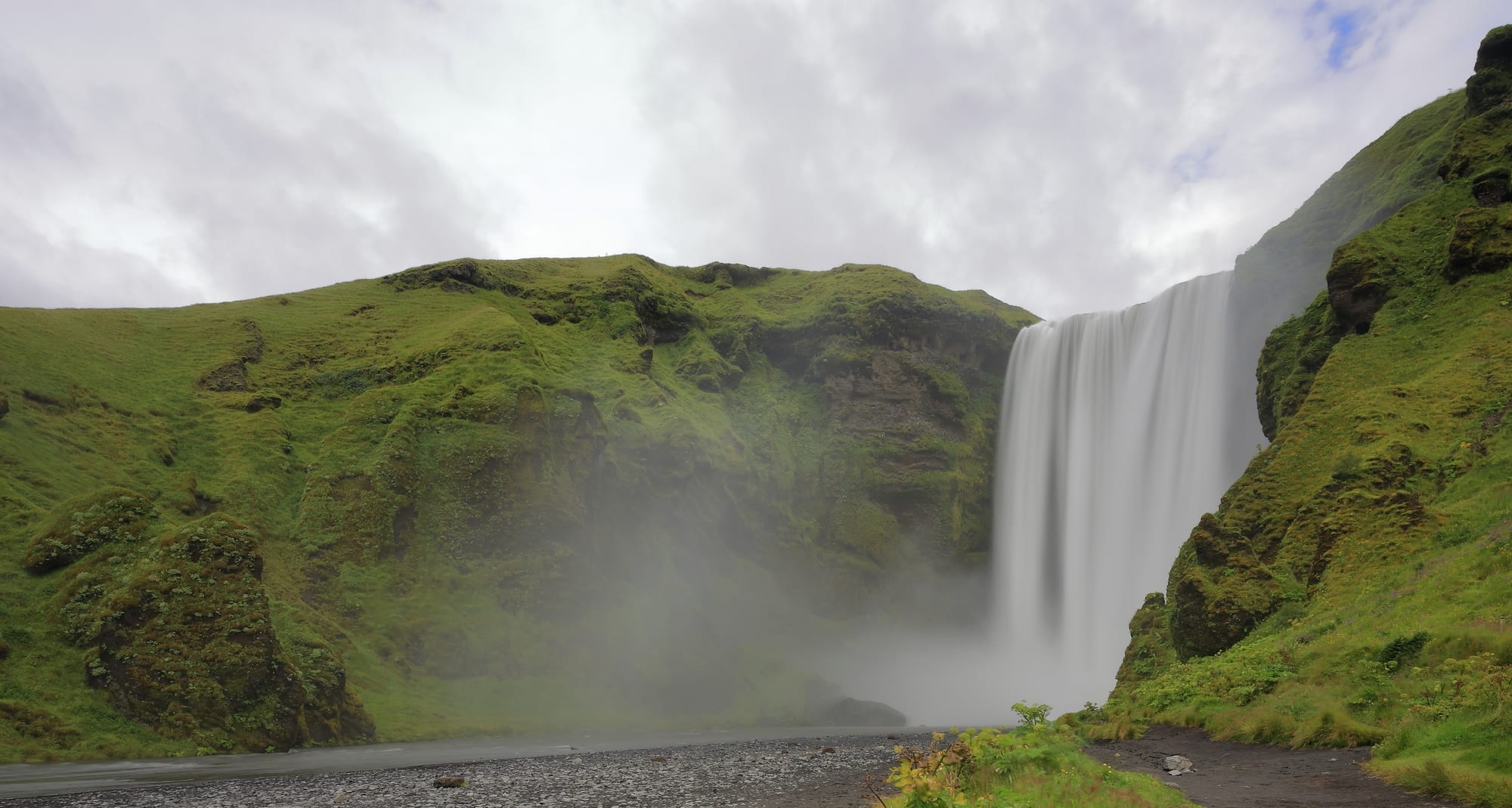 A wide, sheer waterfall dropping from a green clifftop onto a flat plain, with a rainbow