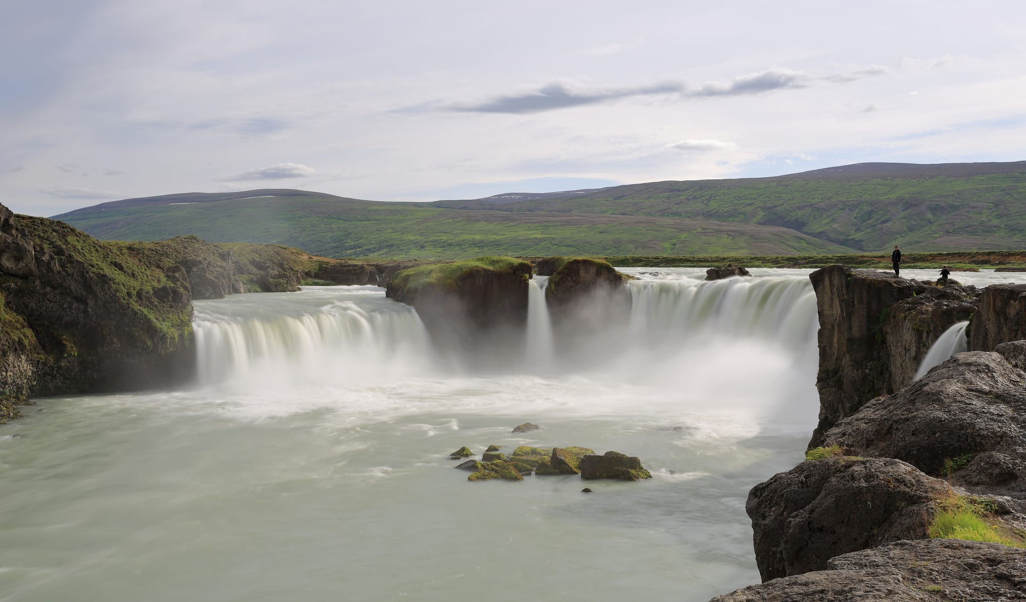 A wide, curved waterfall split by rocks, dropping into a blue river gorge