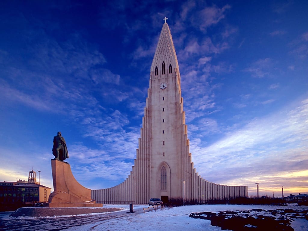 A tall pale concrete church with a stepped, wing-like tower rising over low colourful houses