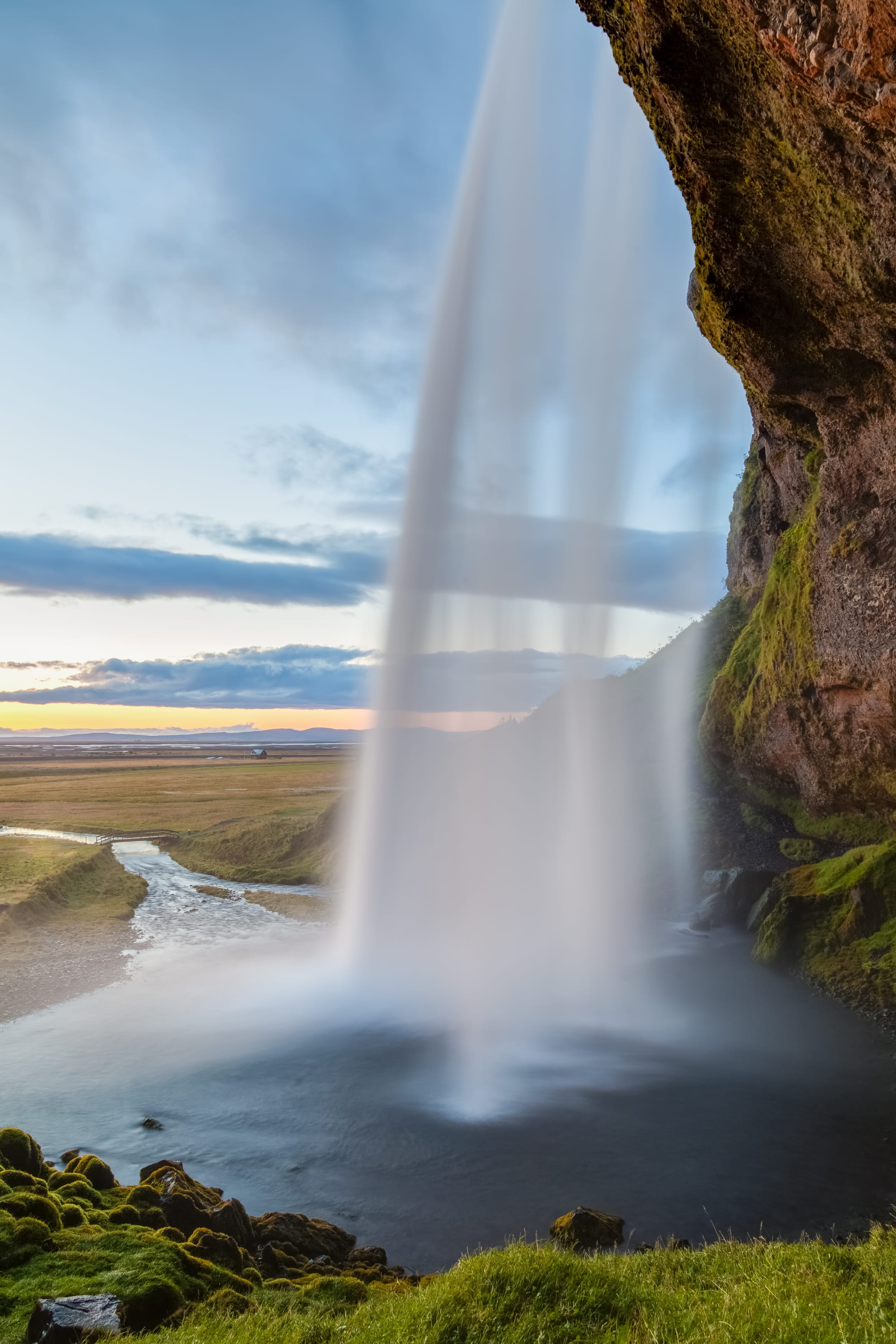 A tall thin waterfall dropping from a green cliff with a footpath running behind the water
