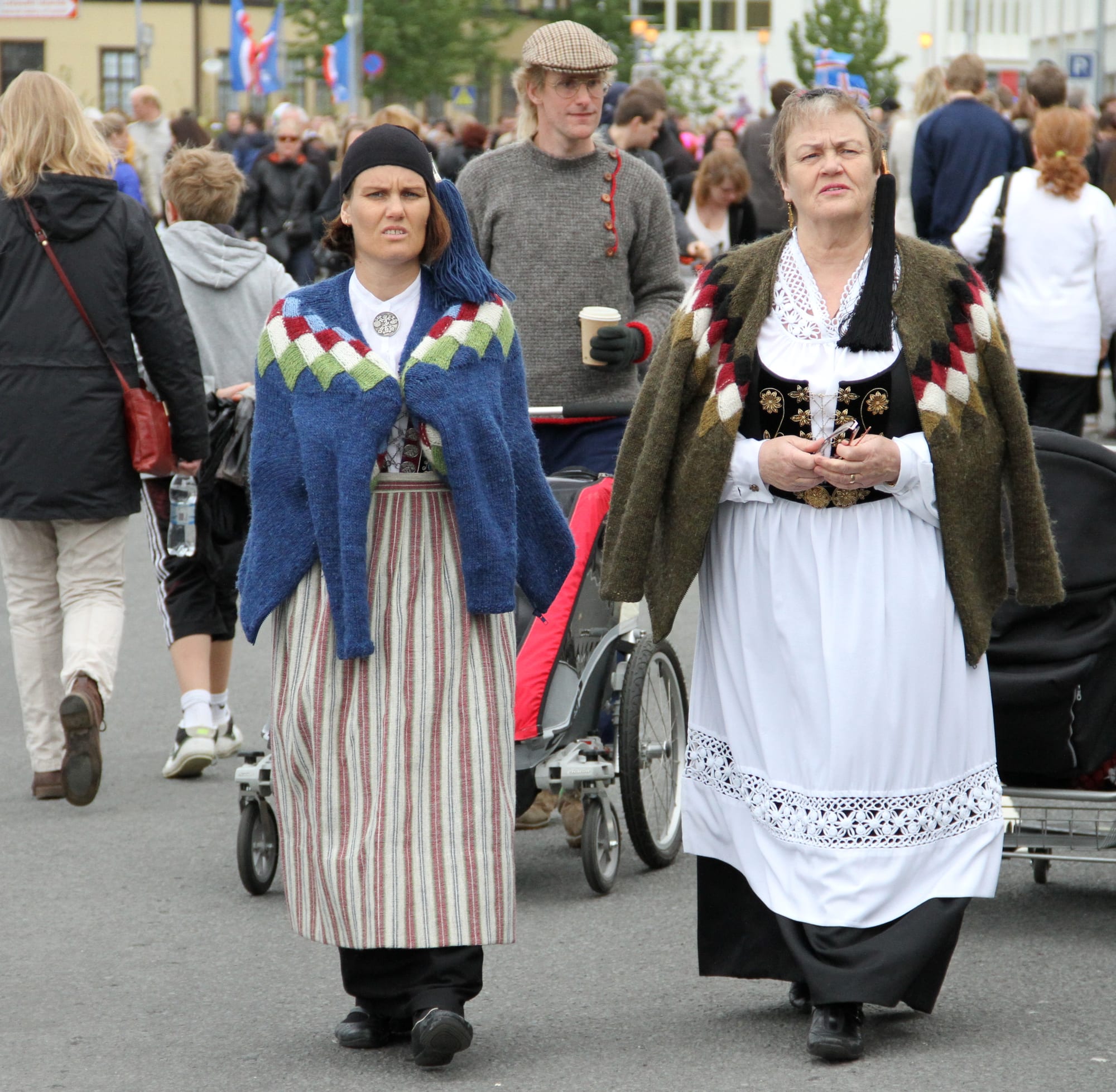 Two women in Icelandic national costume, one with a striped skirt and blue knitted shawl, the other in a white skirt with a black embroidered bodice and green shawl, walk along a crowded street