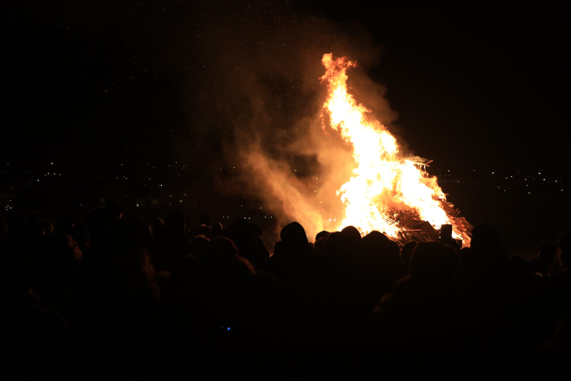 A large bonfire blazes against a dark night sky, with sparks and smoke rising and a crowd silhouetted in the foreground