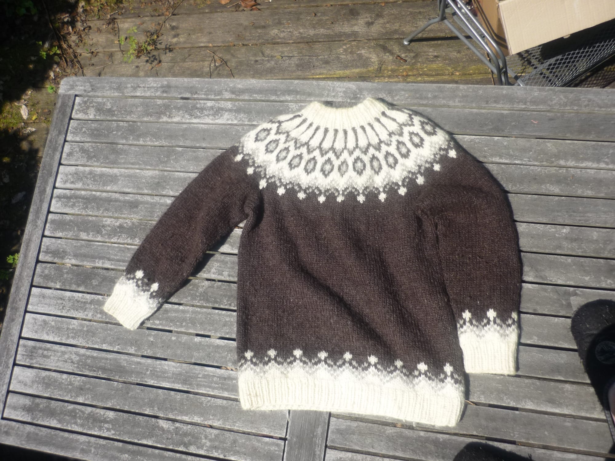 A dark brown hand-knitted wool sweater with a cream patterned yoke and matching cream bands at the cuffs and hem, laid flat on a weathered wooden table