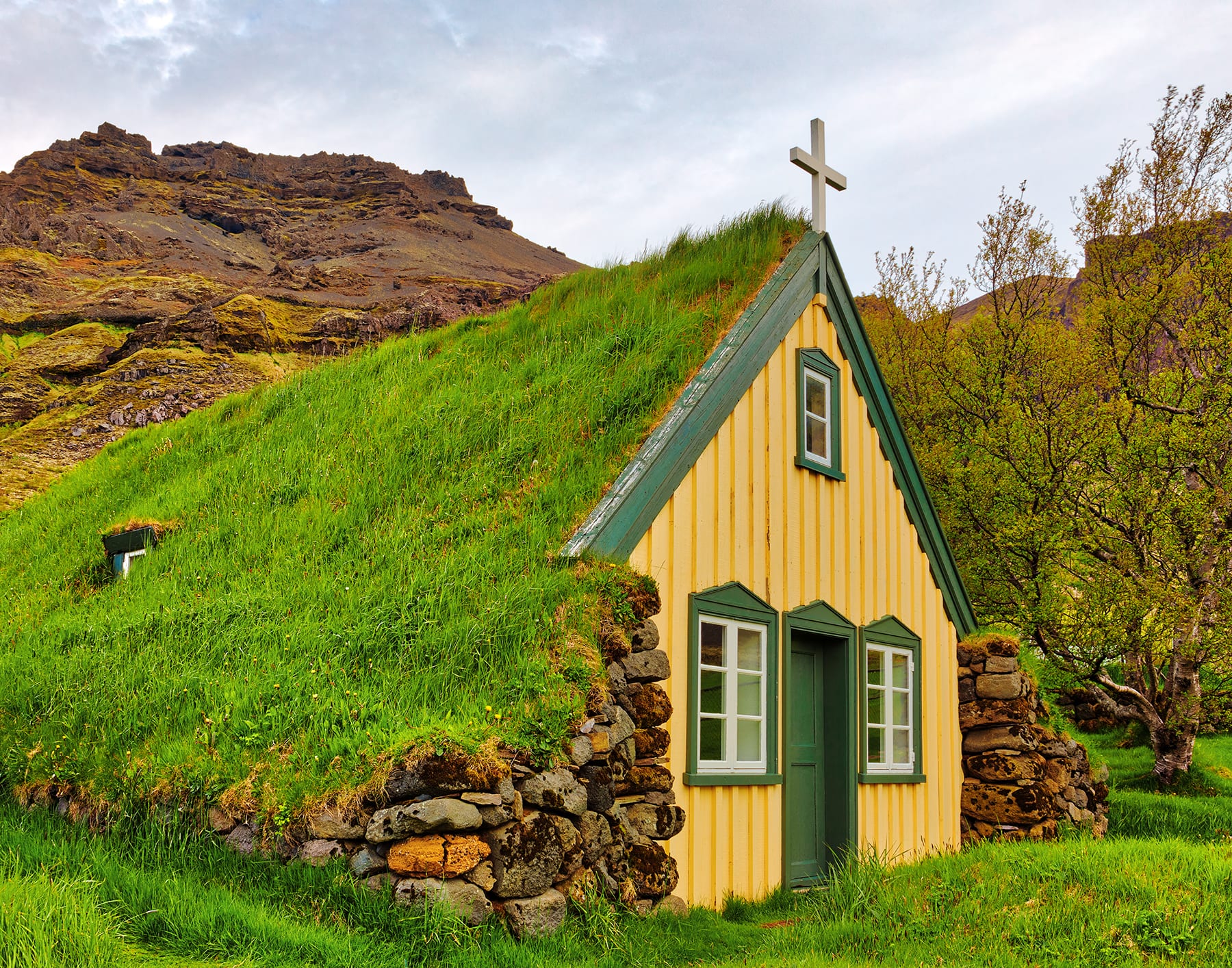 A small church with a steep grass-covered turf roof, a pale yellow wooden gable with green-framed windows and a white cross on top, set against a rocky mountainside